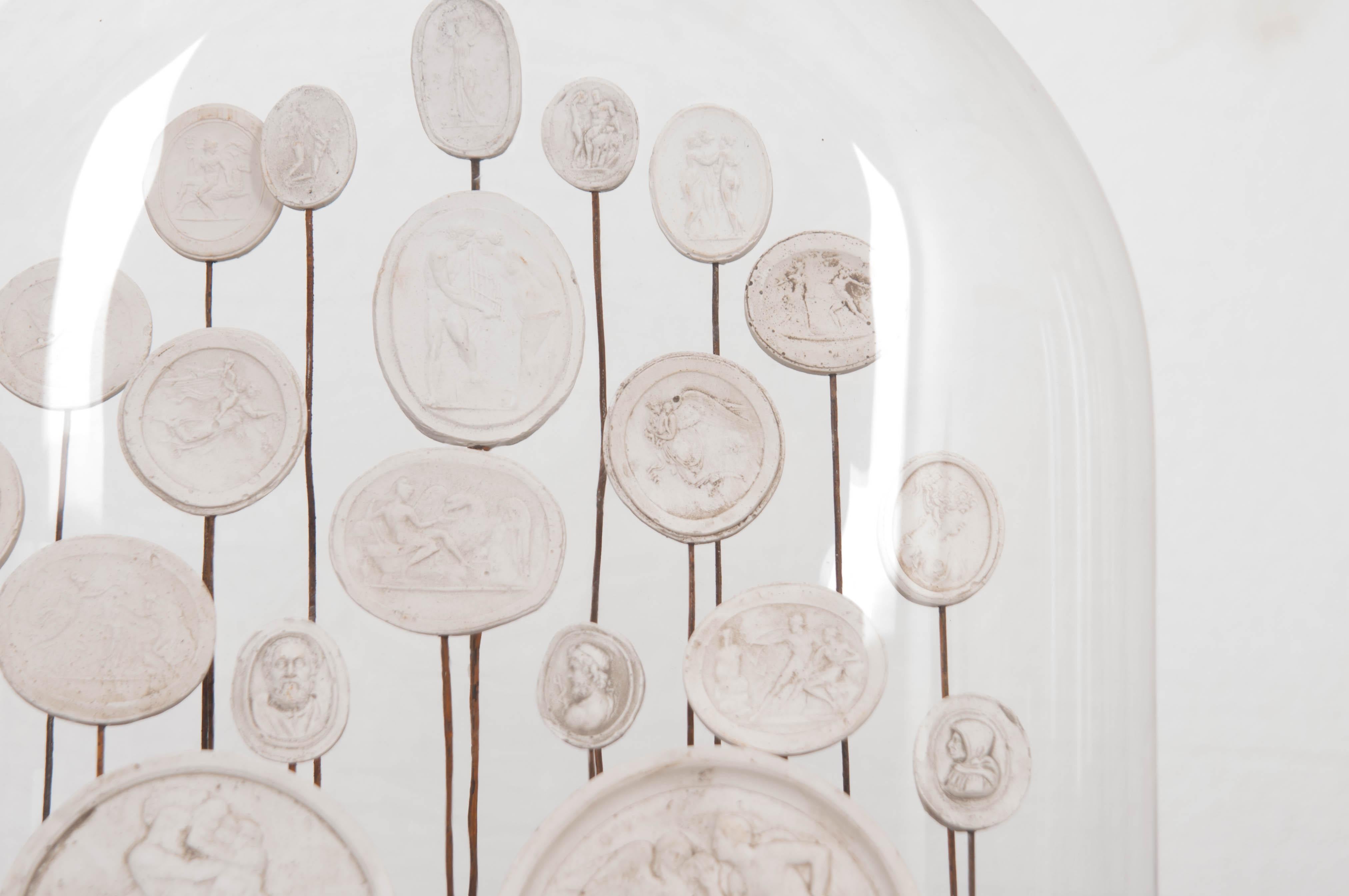 A beautiful assortment of recently-made plaster intaglios set underneath an antique English glass dome cloche. The intaglios have undergone an antiquing process that give them the appearance of age. Intaglios are one of the earliest forms of