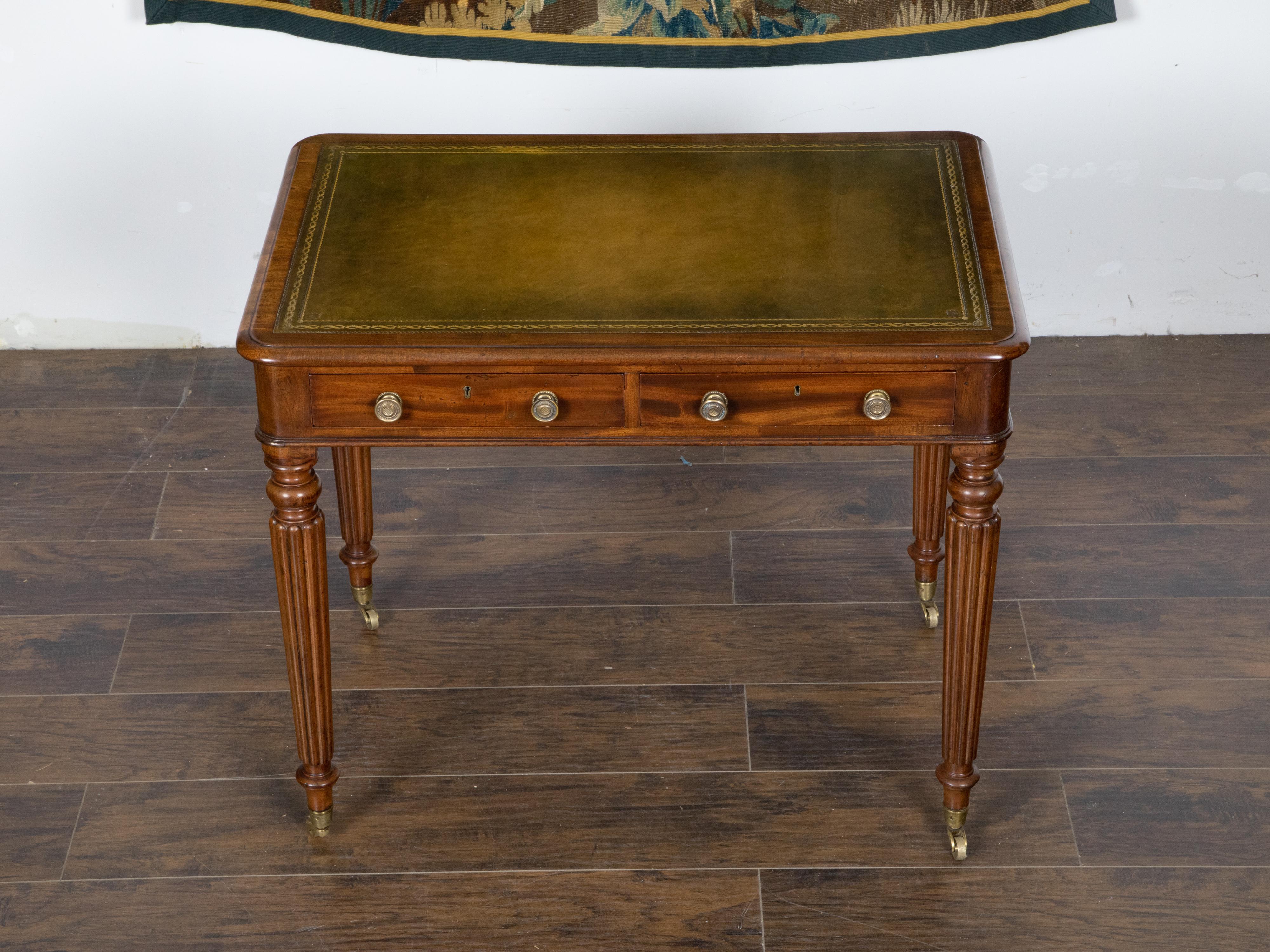An English mahogany writing table from the 19th century, with green leather top, gilt accents, carved reeded legs, two drawers and brass casters. Created in England during the 19th century, this desk features a rectangular top with rounded corners,
