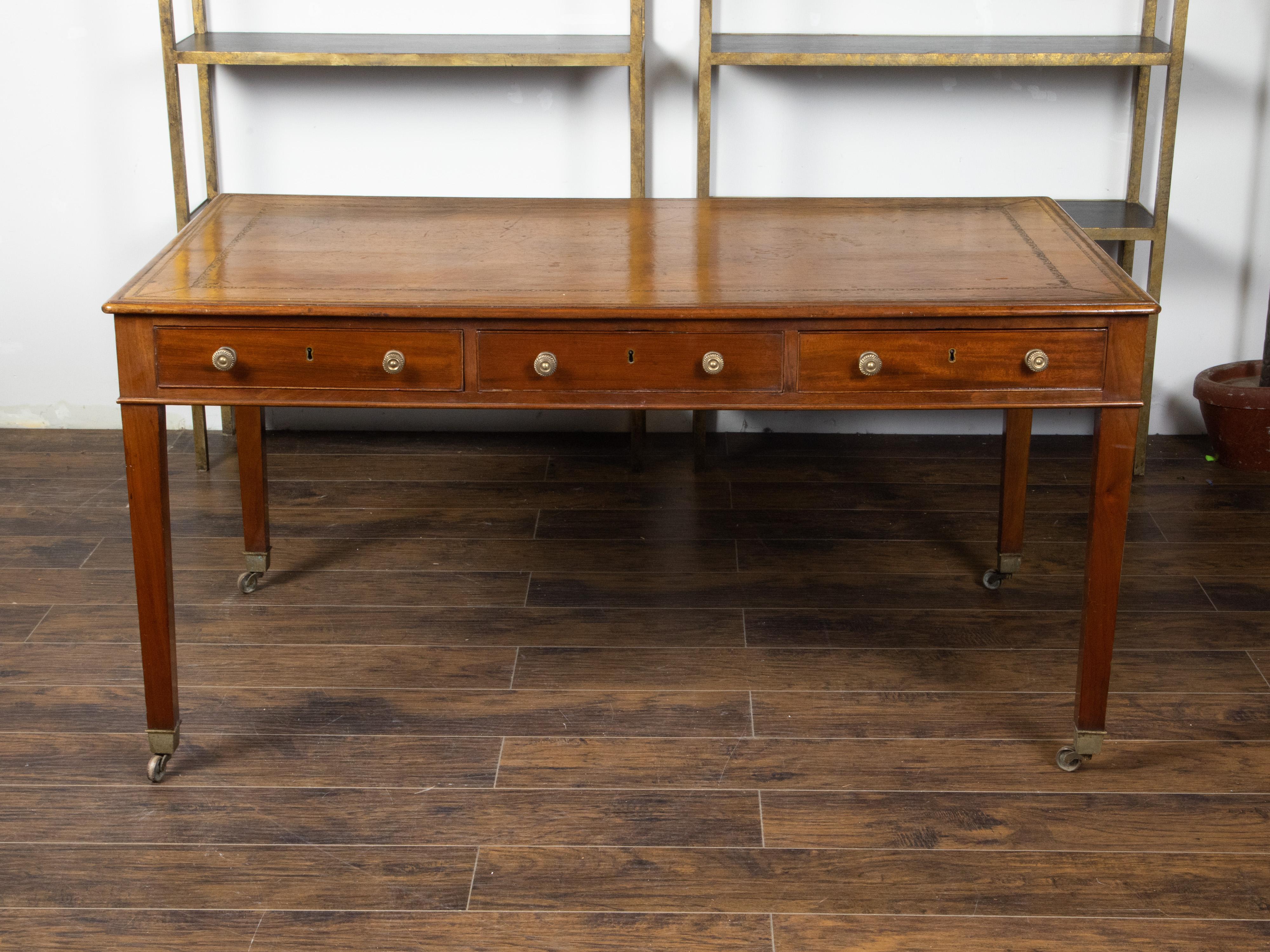 An English mahogany desk from the 19th century, with leather top, three drawers and casters. Created in England during the 19th century, this mahogany desk features a rectangular brown leather top and gilt accents, sitting above three drawers fitted