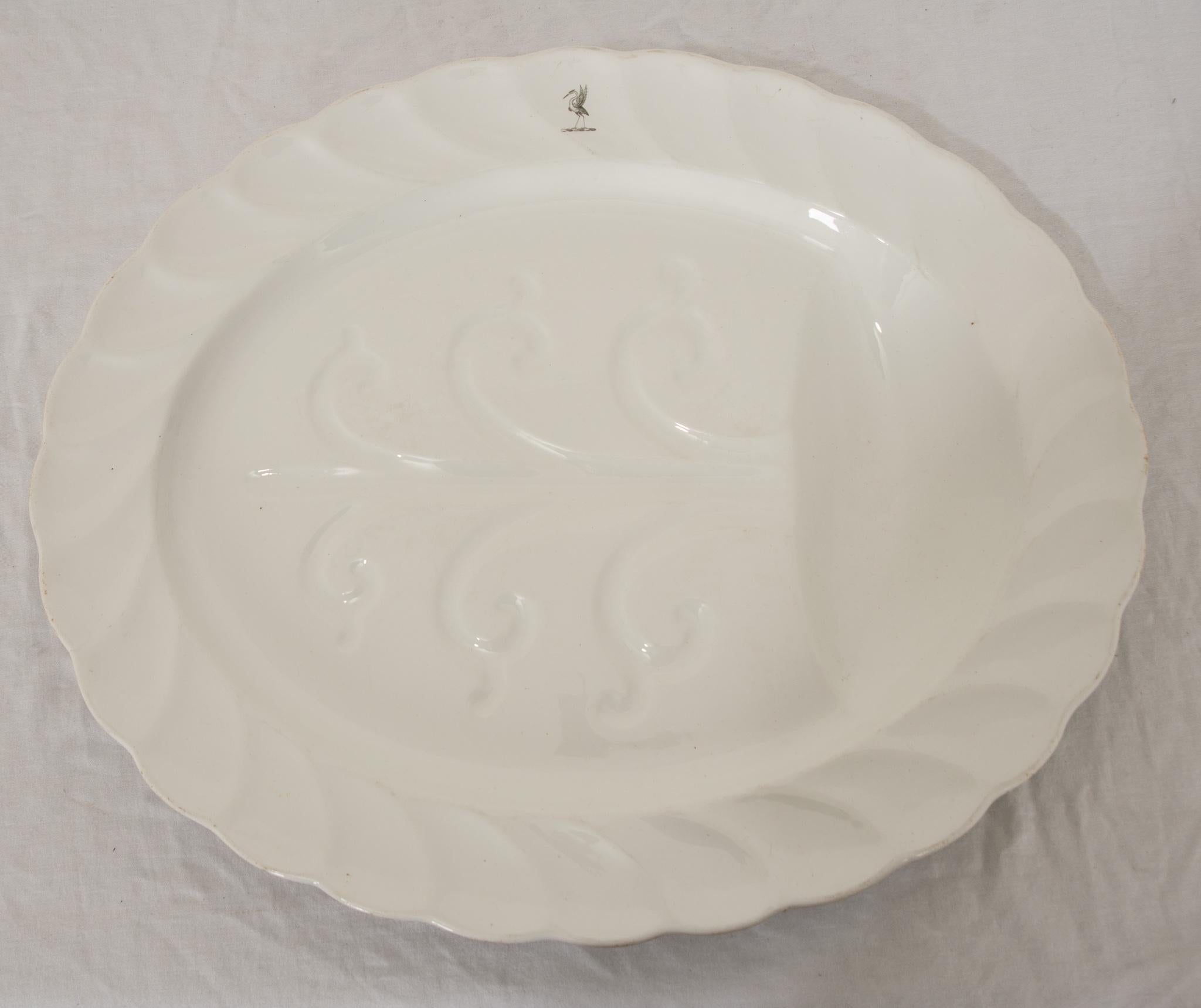 This stately 19th Century English porcelain meat platter features a scalloped rim, embossed deep draining channels, and is replete with basting well at one end.  Raised upon integral feet to base. High gloss glaze overall with a decorative crane on
