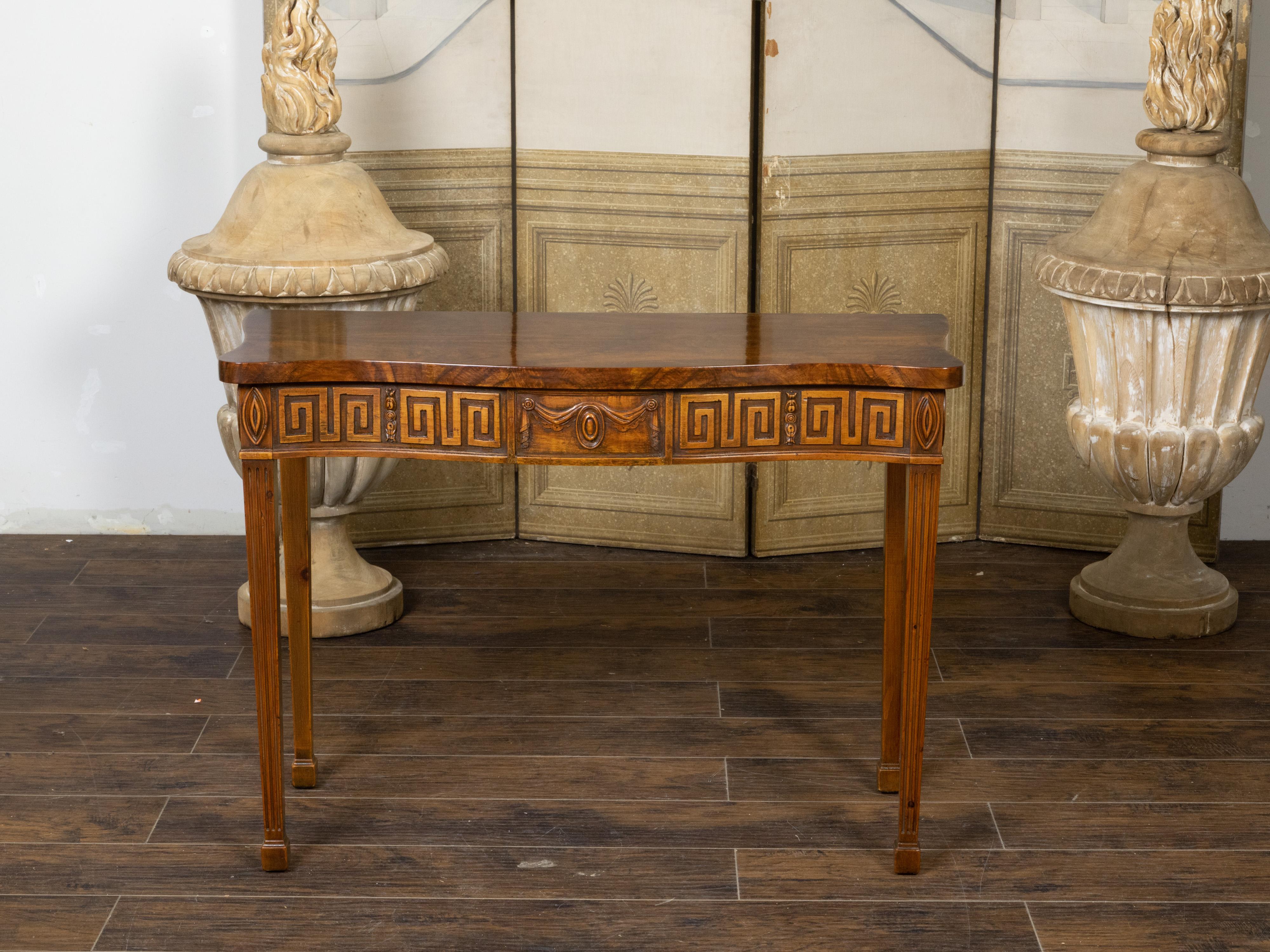 An English Neoclassical Revival mahogany console table from the 19th century, with carved apron depicting a Greek Key frieze and a draped oval medallion. Created in England during the 19th century, this console table features a serpentine top