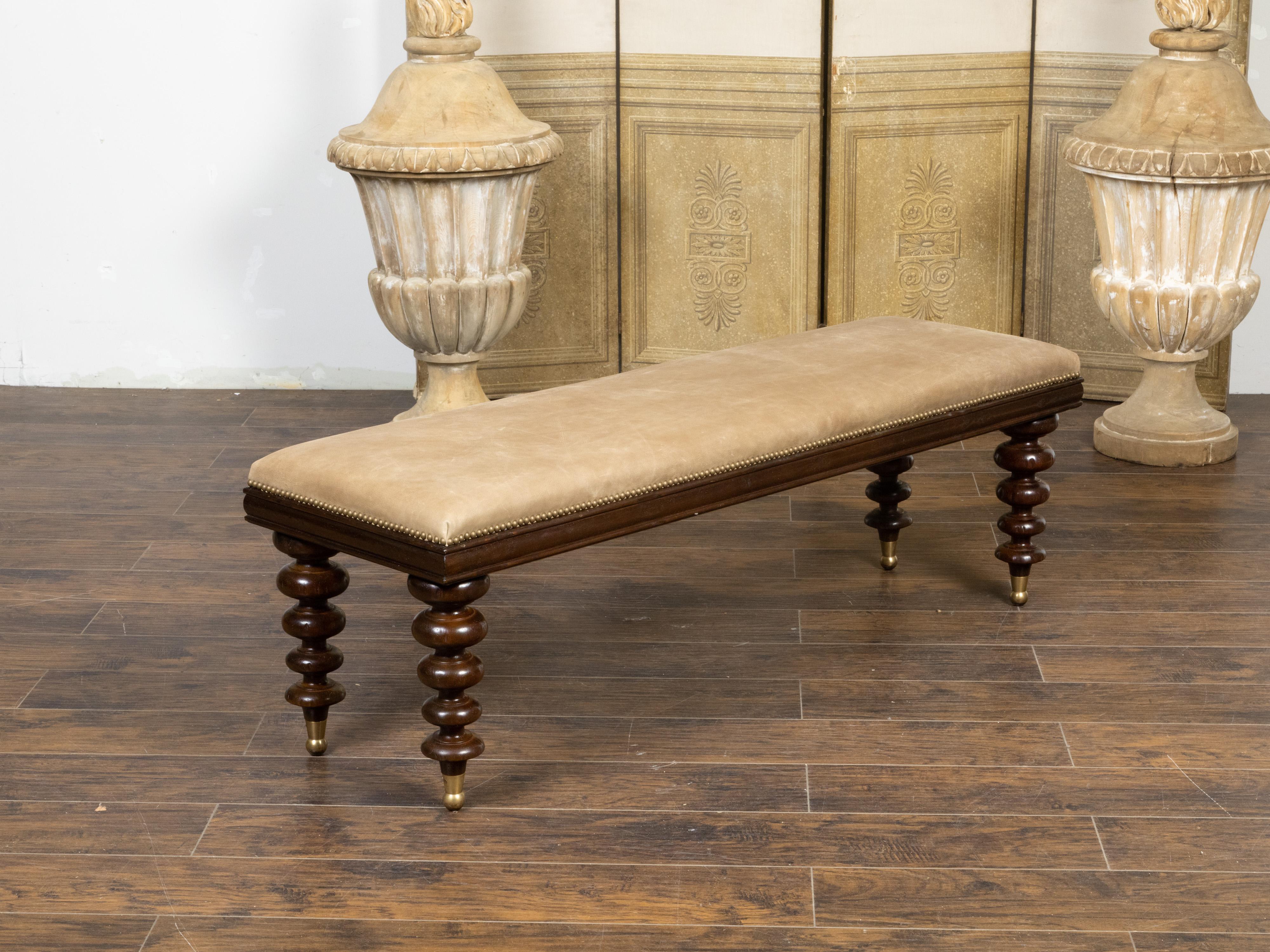 An English oak bench from the 19th century, with spool turned legs, leather upholstery, nailhead trim and brass feet. Created in England during the 19th century, this oak bench features a rectangular seat covered with a camel color leather fabric