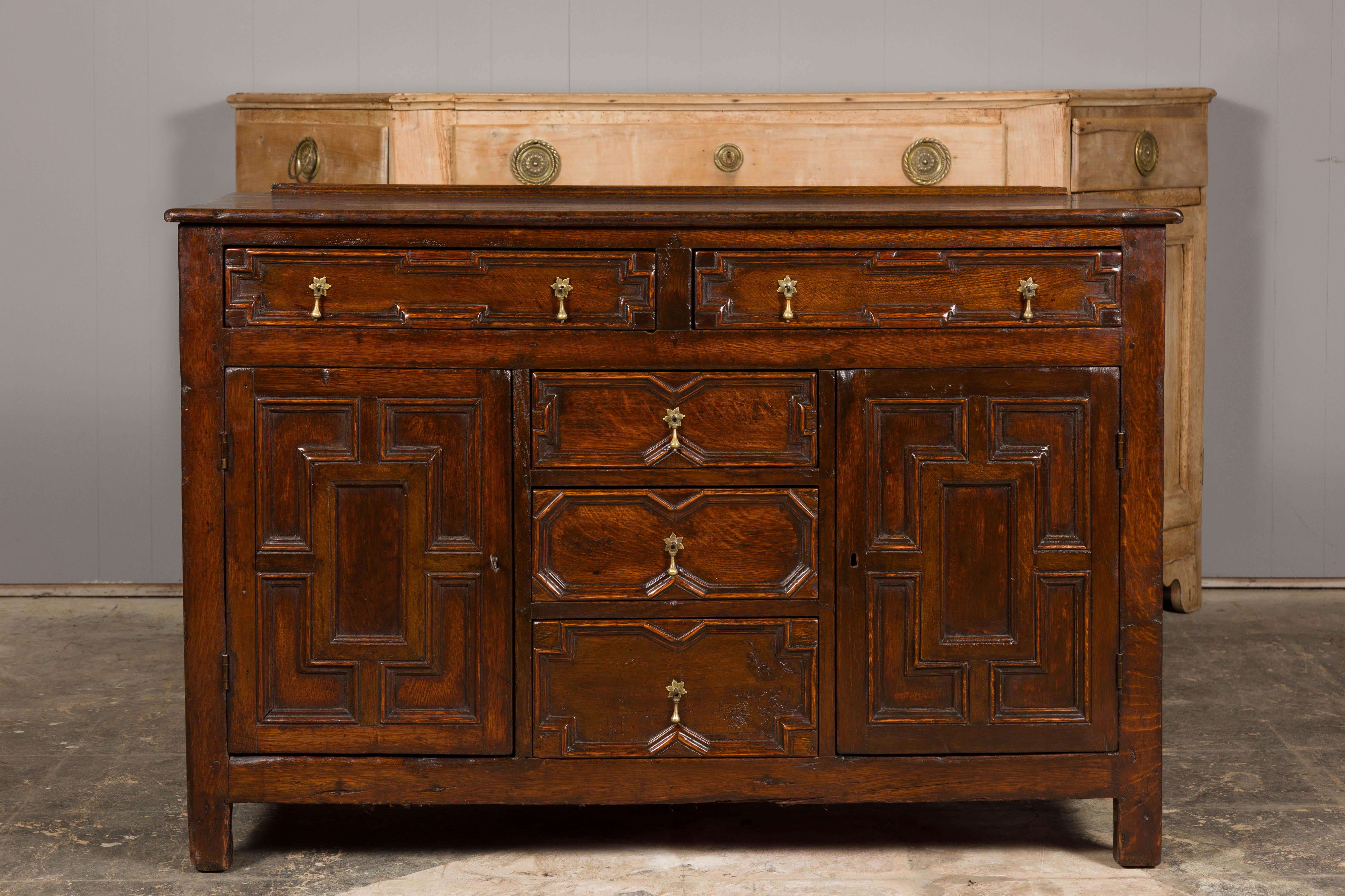 An English oak dresser base from the 19th century with geometric front, five drawers, two doors and brass hardware. Immerse in the rustic charm and time-honored craftsmanship of this 19th century English oak dresser base, a delightful convergence of