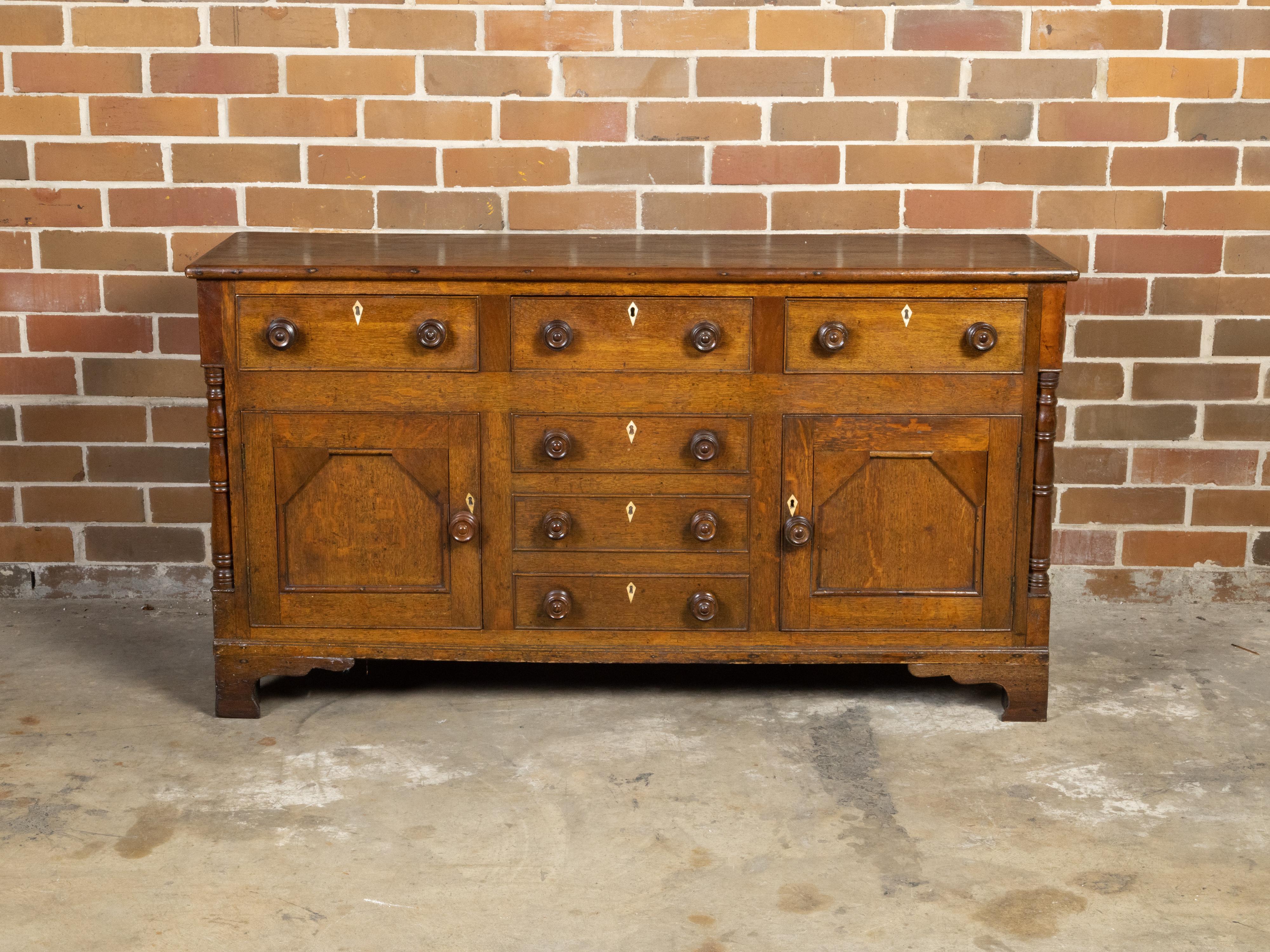 An English oak dresser base from the 19th century with six drawers, two doors, bone inlaid escutcheons and thin columns. Created in England during the 19th century, this oak dresser base features a rectangular planked top sitting above a perfectly