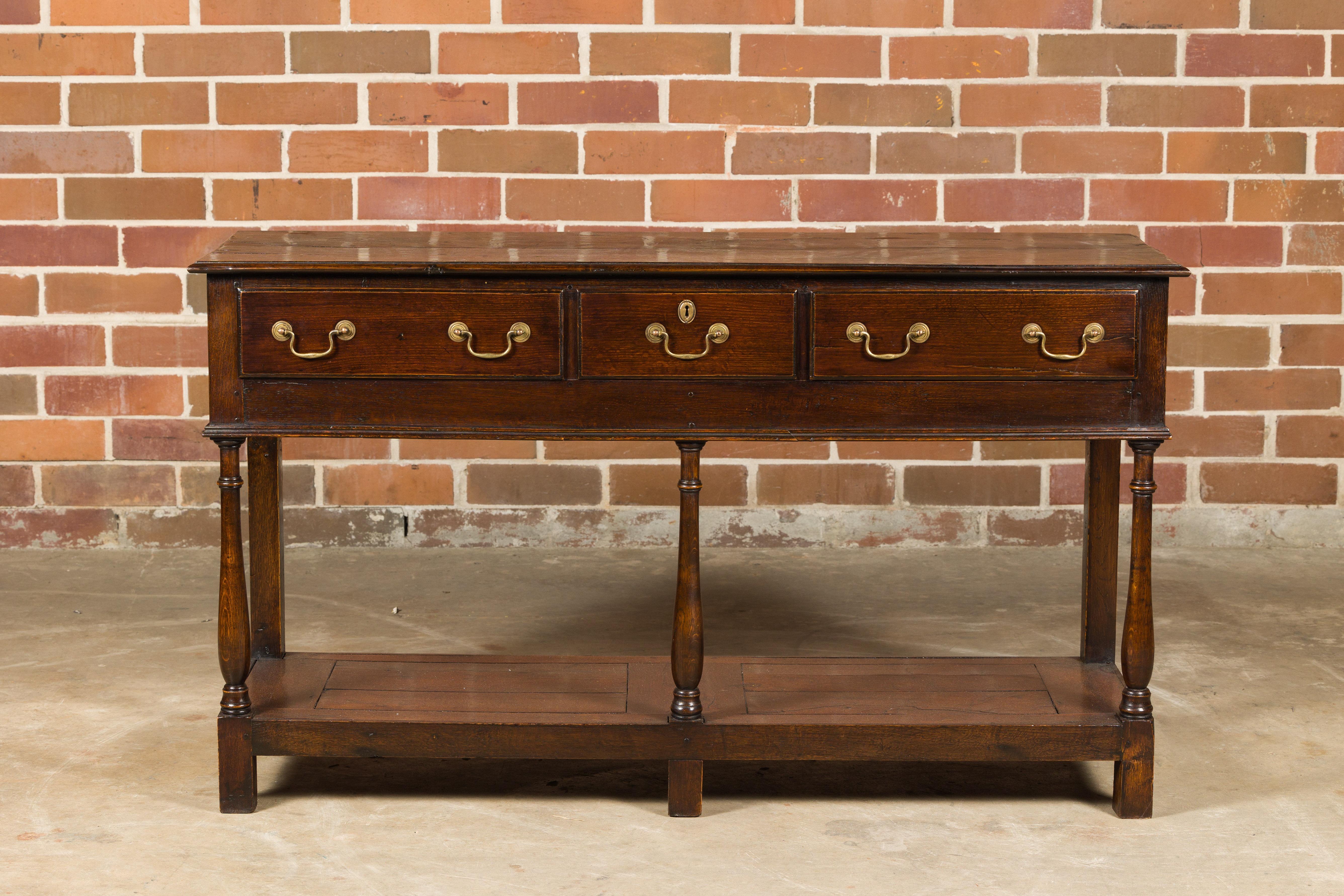 An English oak dresser base from the 19th century with three drawers, low shelf and turned baluster legs. This 19th-century English oak dresser base is a testament to enduring craftsmanship and timeless beauty. Its three drawers, adorned with brass