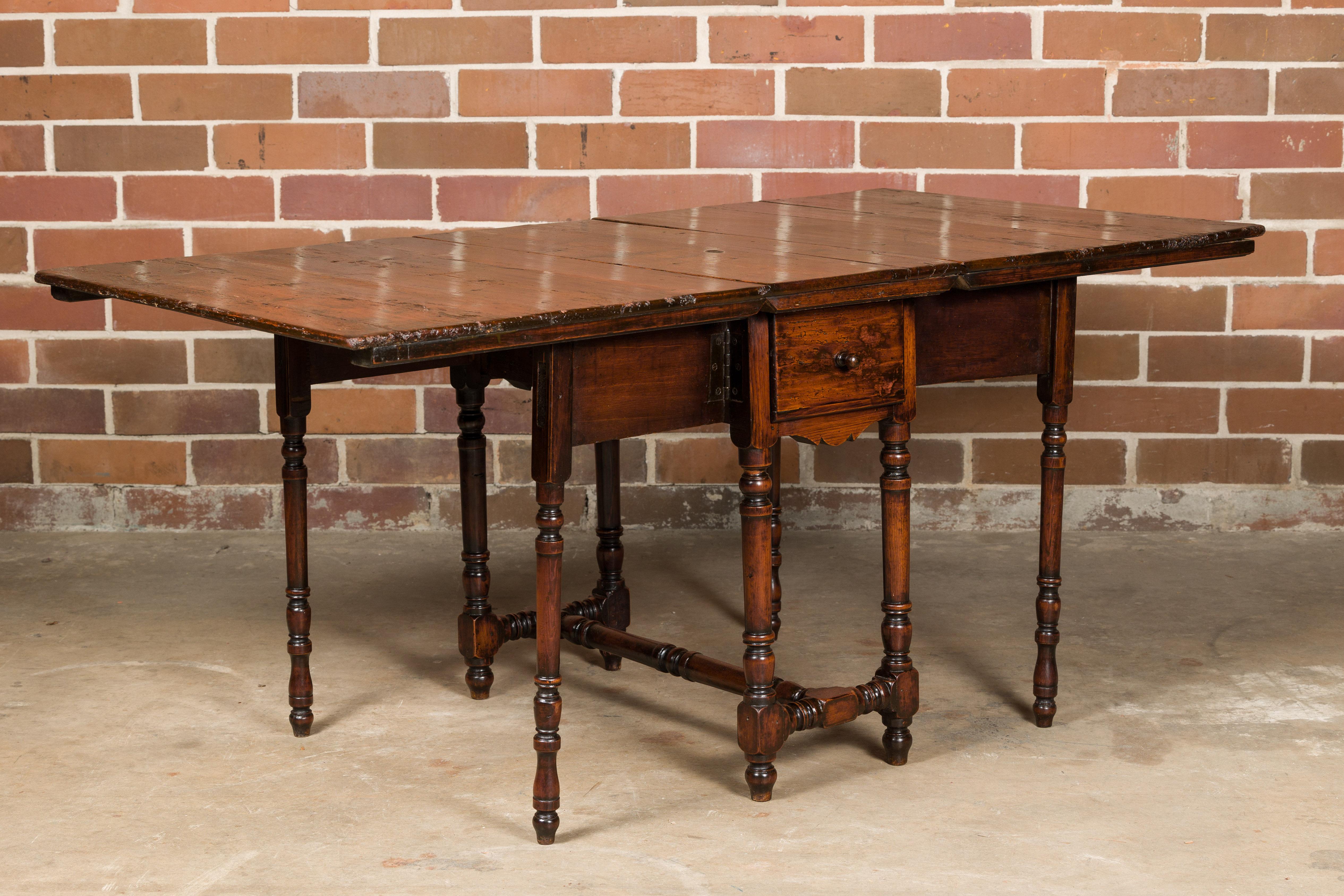 An English oak drop leaf table from the 19th century with slender turned legs, two lateral drawers and H-Form cross stretcher. This English oak drop-leaf table from the 19th century is a splendid example of functional elegance. Crafted with an