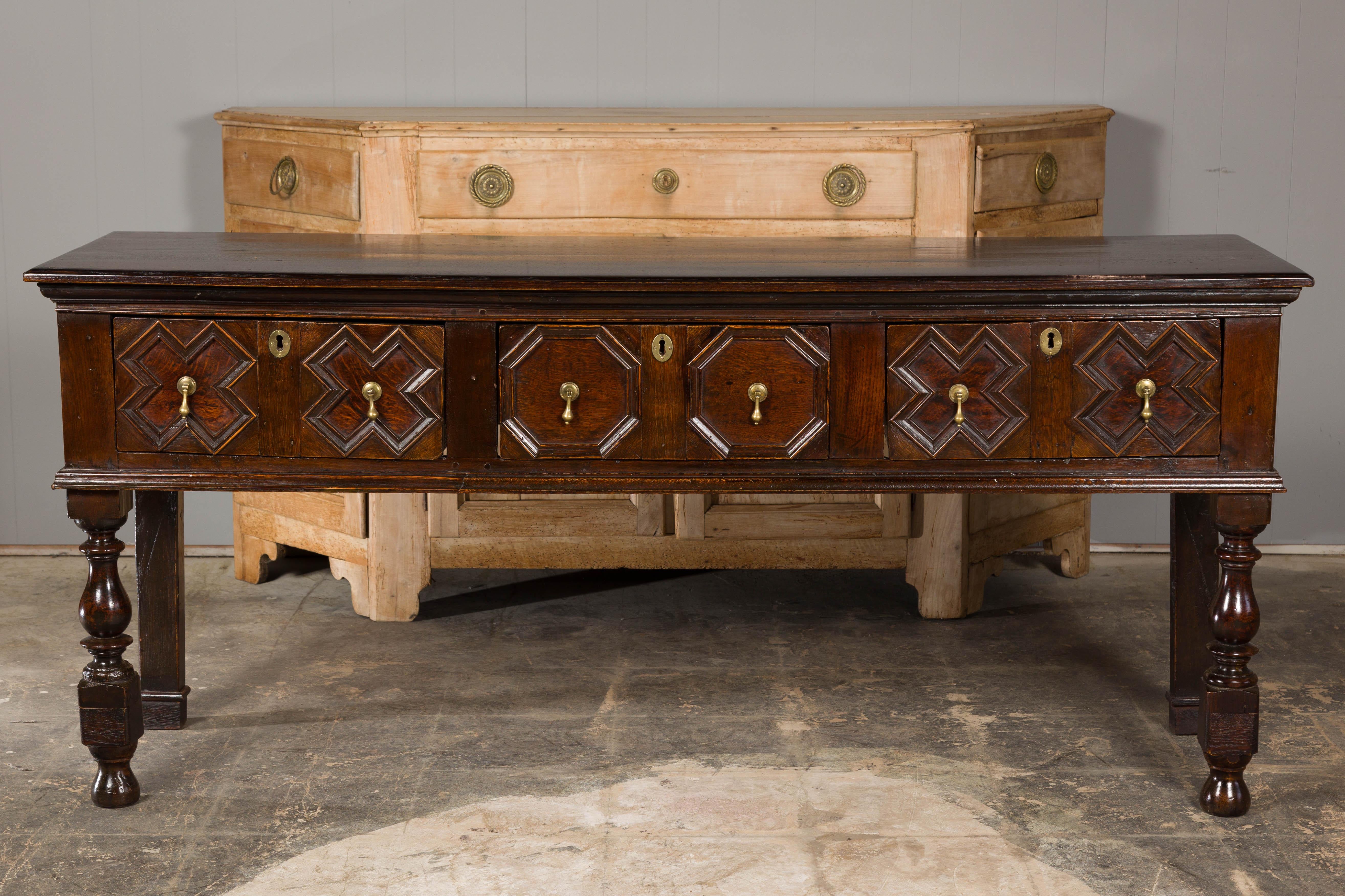 An English oak sideboard from the 19th century with geometric front and turned baluster legs. Enthrall your living space with the charm and regal allure of this 19th-century English oak sideboard, an emblem of craftsmanship and tradition. Cradled by
