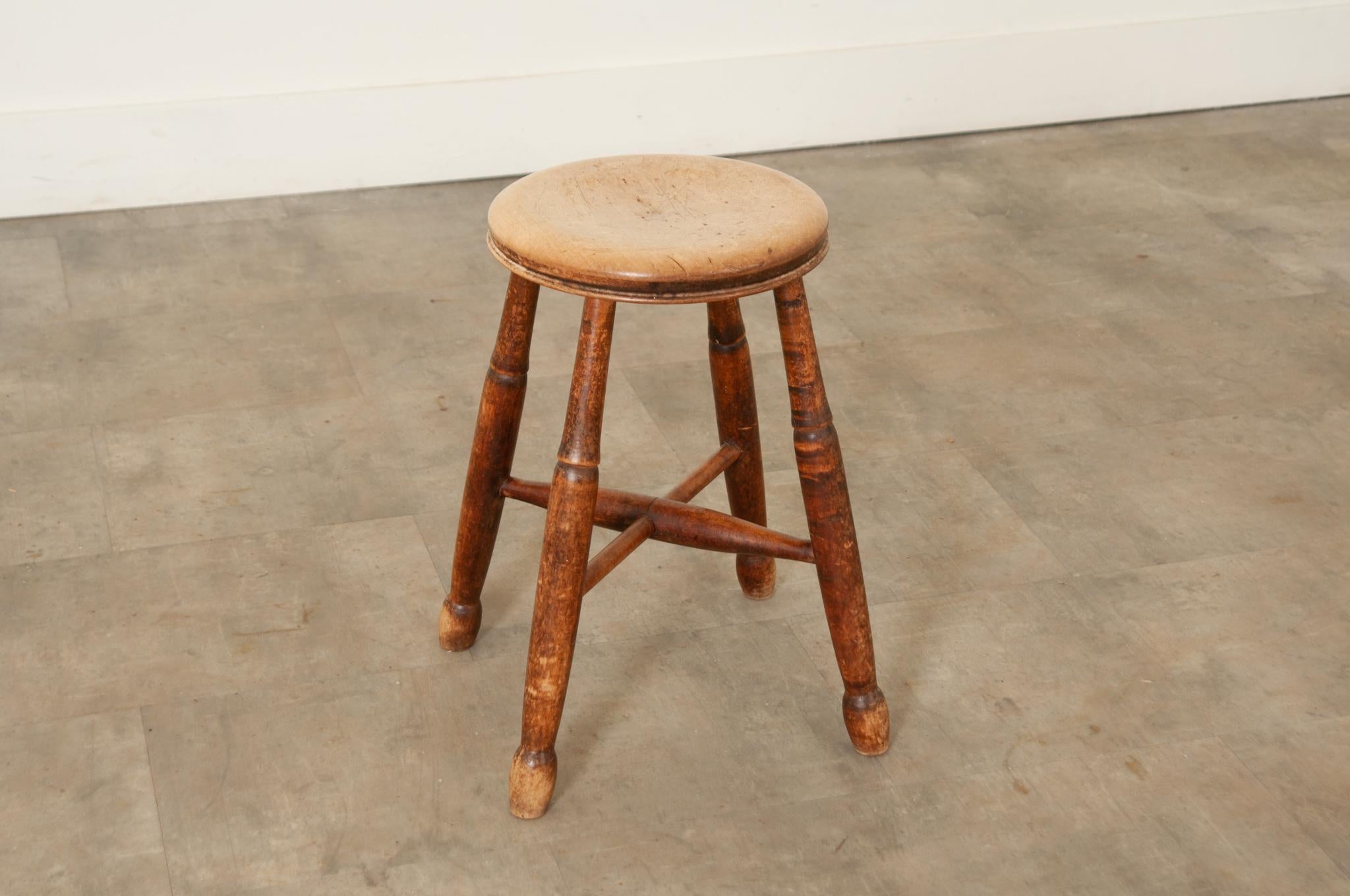 A Victorian English farmhouse stool handcrafted in oak in England circa 1840 featuring a smooth molded and trimmed round seat with comfortable recessed center atop four banded and turned splayed legs joined with a sturdy turned stretcher for extra