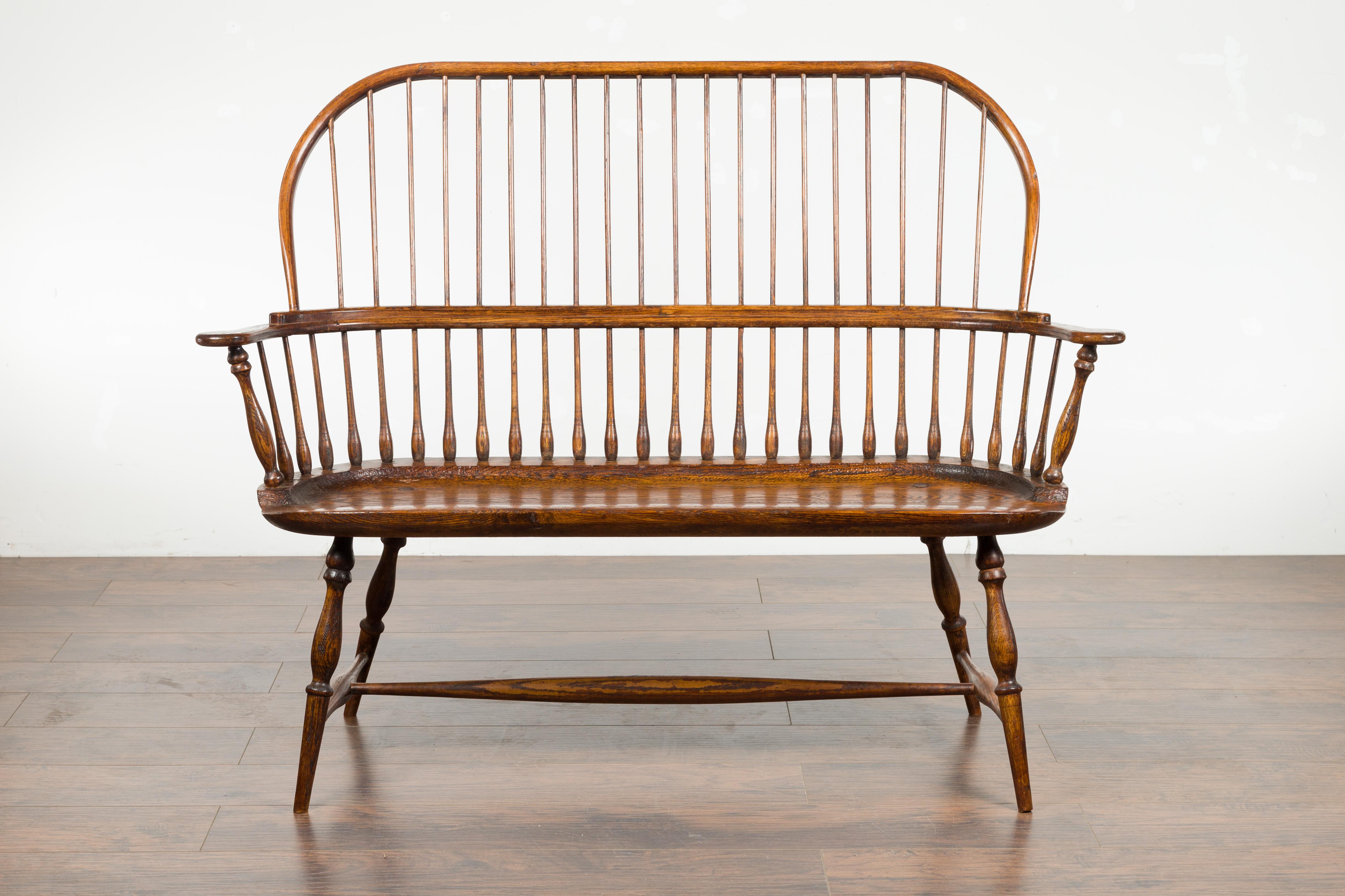 An English oak Windsor bench from the 19th century, with spindle accents and cross stretcher. Created in England during the 19th century, this Windsor bench features a slatted back with spindle motifs in the lower section, sitting on a wooden seat
