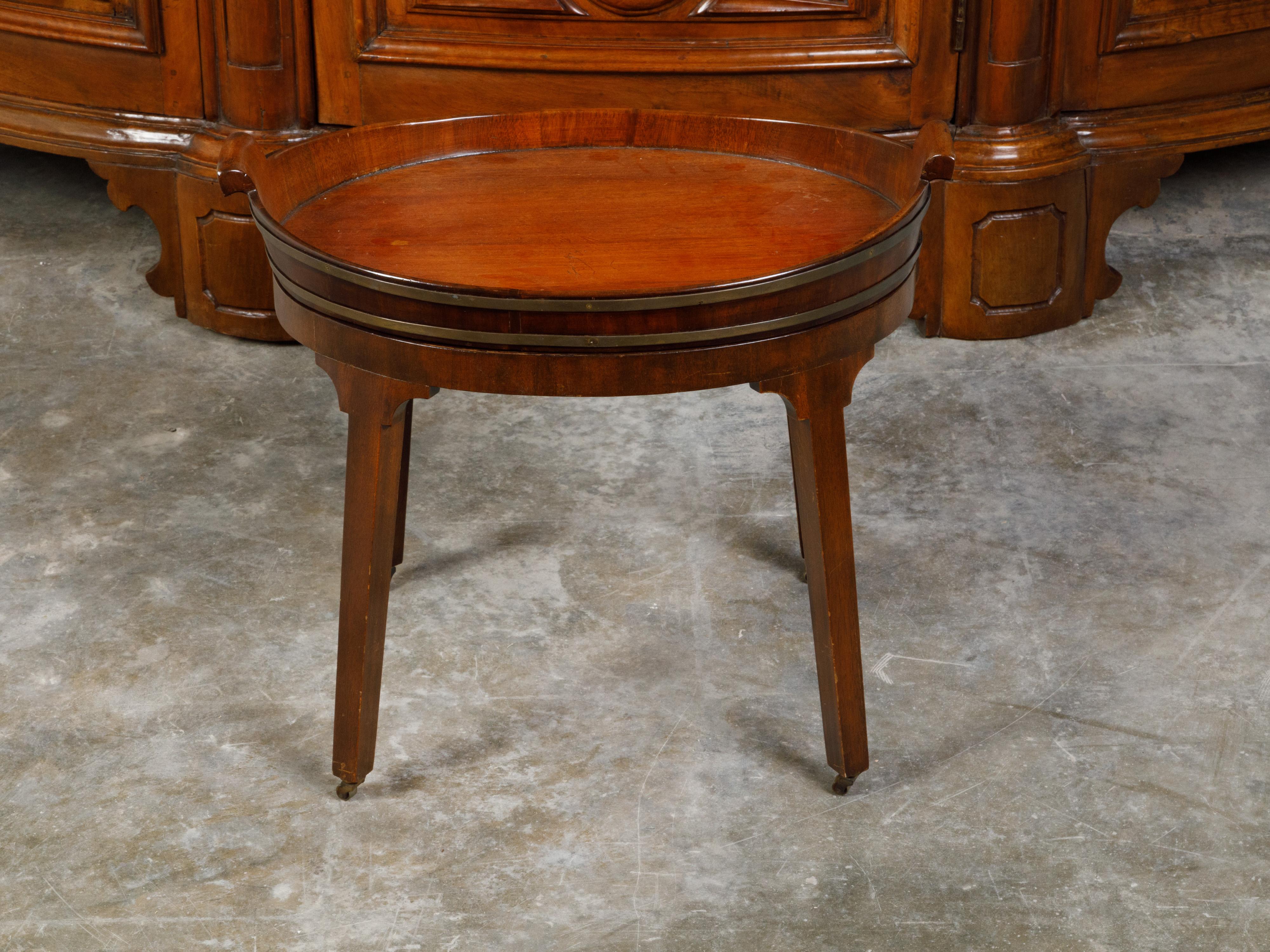 An English mahogany tray top side table from the 19th century, with splaying legs and casters. Created in England during the 19th century, this small side table features an oval tray top with petite out-scrolling handles, accented with brass braces.