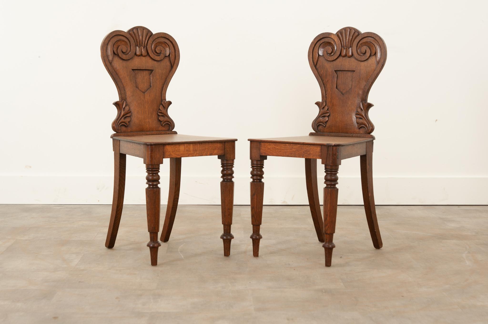 A pair of finely crafted solid oak English hall chairs from the 19th century. The shapely back showcases scrolling and shell inspired hand-carved elements at the top, a recessed shield form in the center, and acanthus leaves. Sturdy and level, they