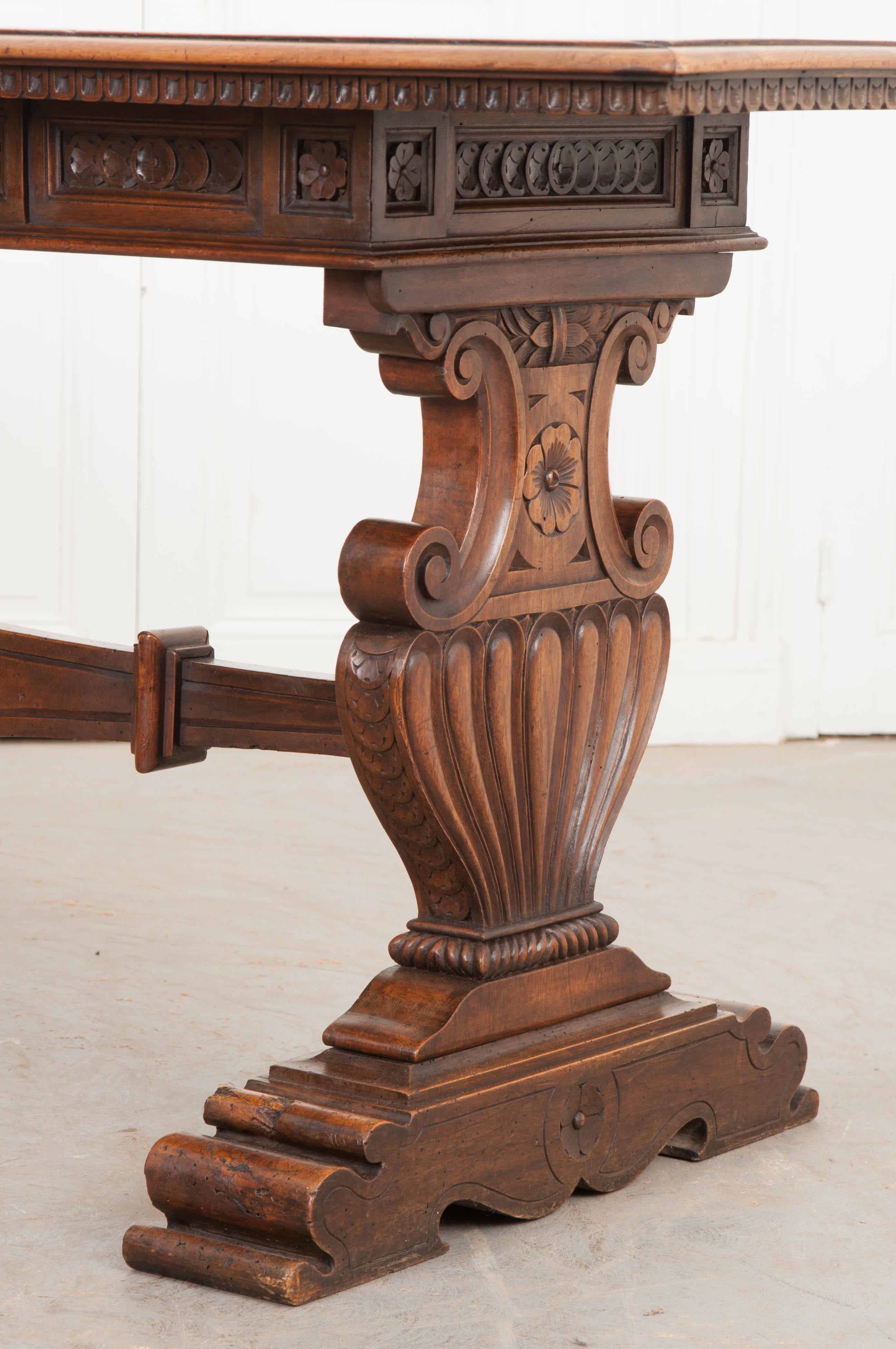 This highly-carved Regency Classicism library table was created in 1830s England and features an elongated rectangular surface with a stepped and dentillated lower edge, atop an apron carved in an “encircled flower head” motif and outfitted with a