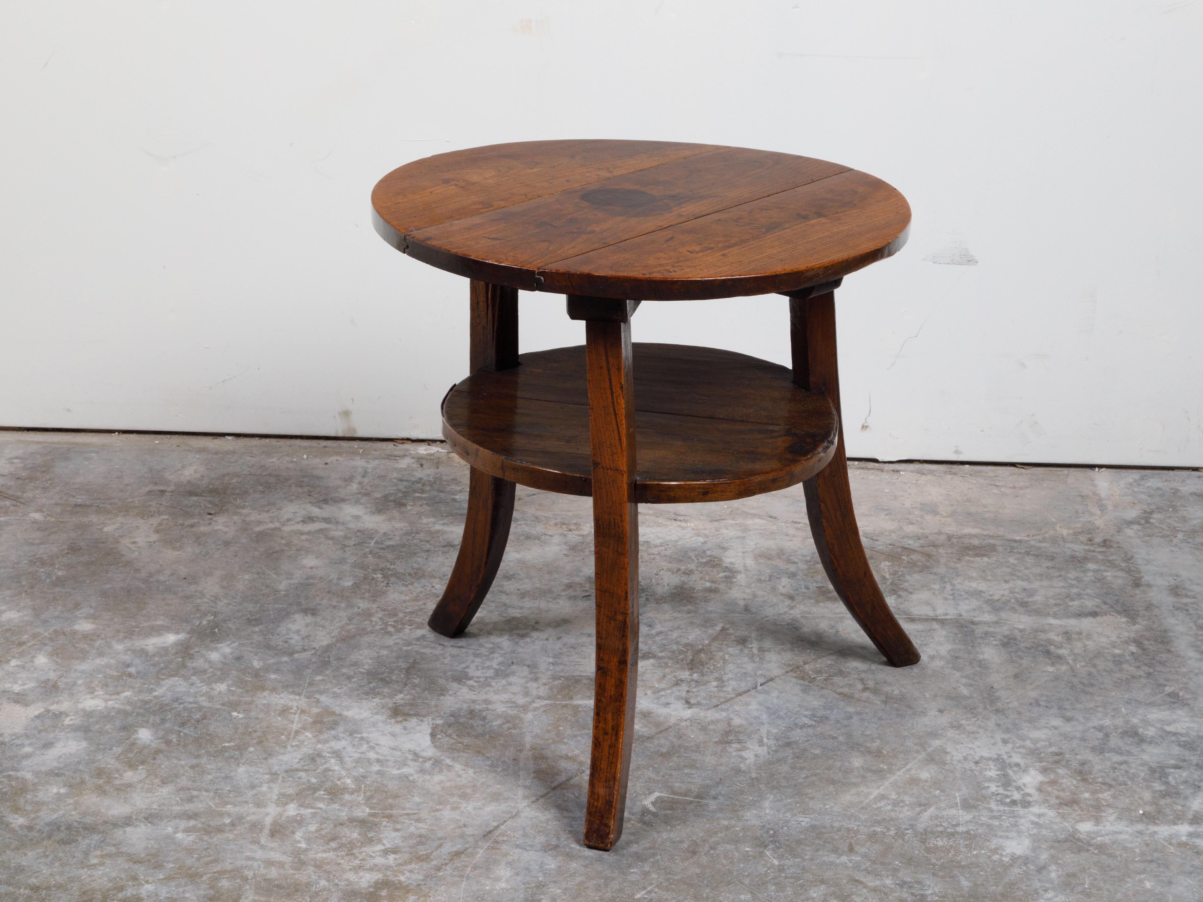 An English walnut side table from the 19th century, with three saber legs, round top and dark patina. Created in England during the 19th century, this walnut table features a circular planked top resting above three saber legs securing a lower shelf