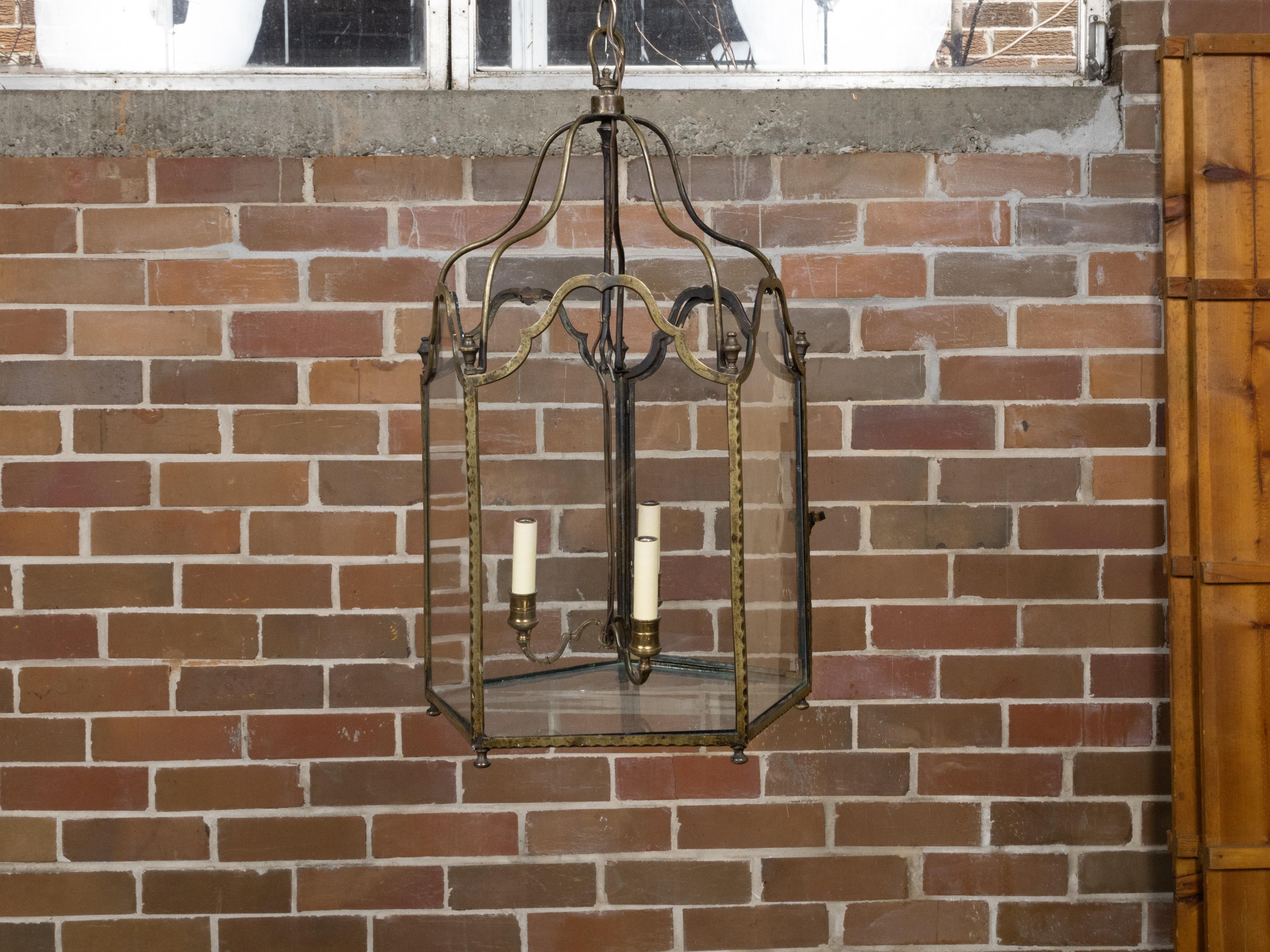 Farol inglés de bronce de tres luces del siglo XIX con paneles de cristal y detalles arqueados. Esta exquisita linterna inglesa de bronce de tres luces del siglo XIX rezuma elegancia atemporal y maestría artesanal. Envuelta en cinco paneles de