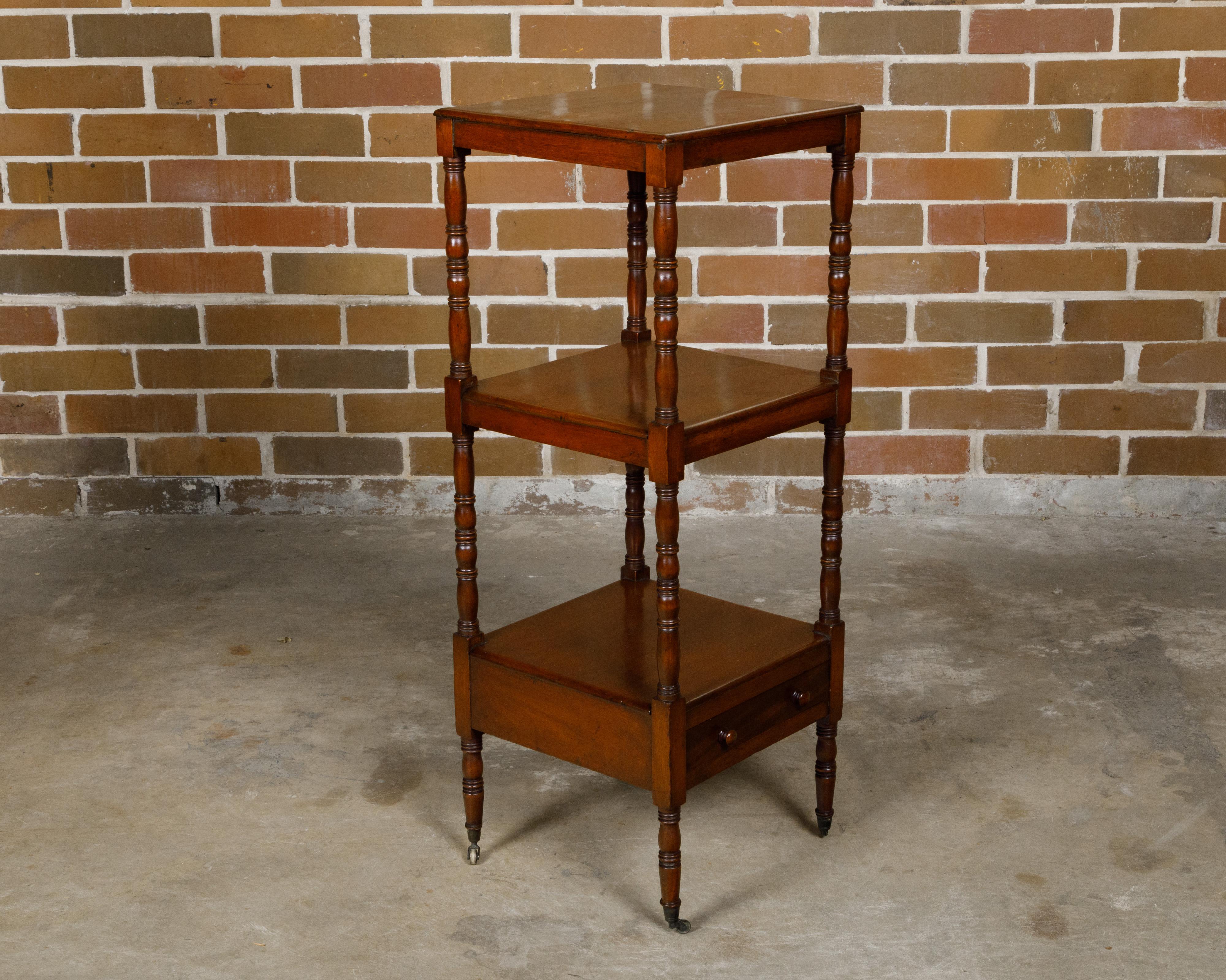 An English mahogany three-tiered étagère from the 19th century with lower drawer, turned supports and casters. This exquisite English mahogany étagère, hailing from the 19th century, elegantly stands on casters, offering both mobility and grace. It