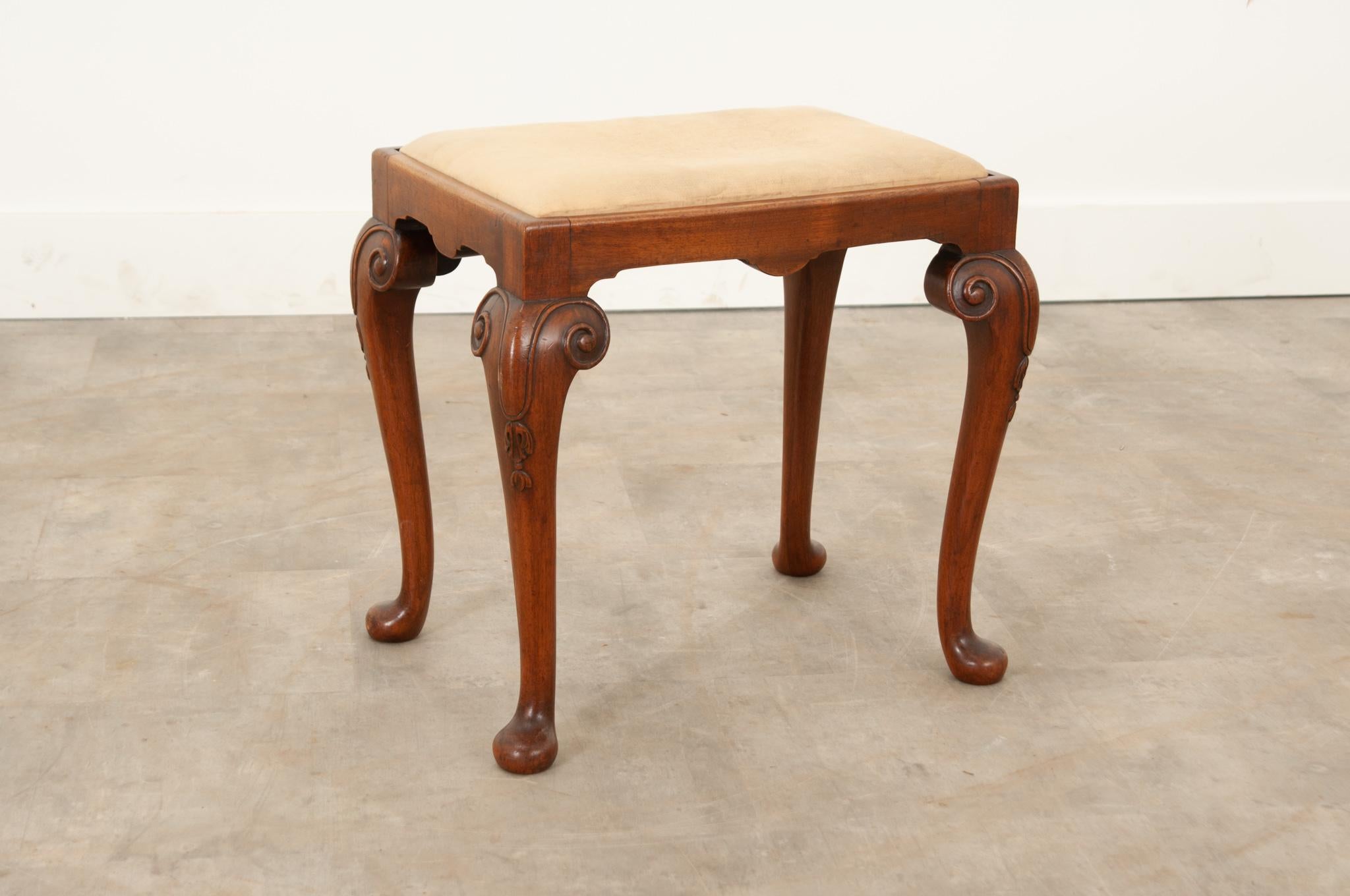 A darling English Regency style stool circa 1880. The solid walnut frame showcases swirling and folate carved designs on the legs and finishes with spoon feet. Upholstered in worn champagne toned fabric and backed with burlap, the removable cushion