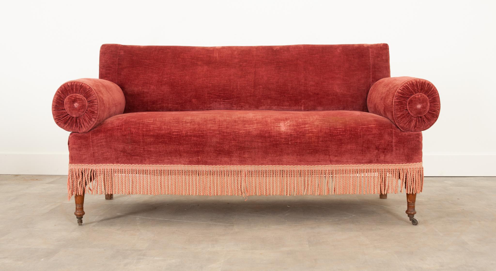 Add a unique pop of color and texture to your interior with this fabulous settee. In wonderful antique condition, the sturdy frame is upholstered in a soft red velvet with a twisted fringe trim. This is an incredibly versatile piece. The arms have