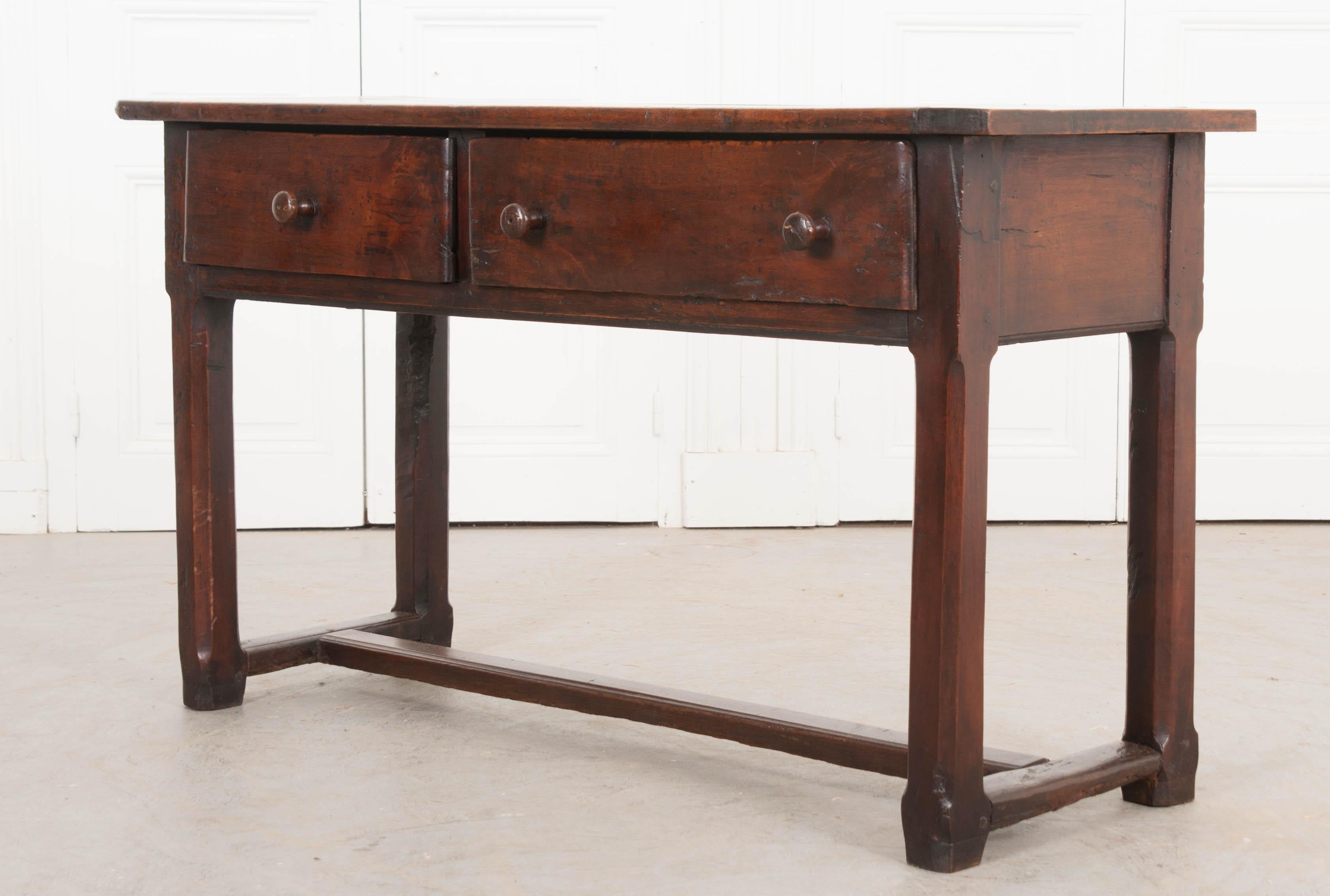 This handsome trestled walnut server, is from 1830s England and has developed a beautiful patina over time. The plank surface rests upon an apron outfitted with two drawers with turned knobs – one large drawer, ideal for linens, and one smaller