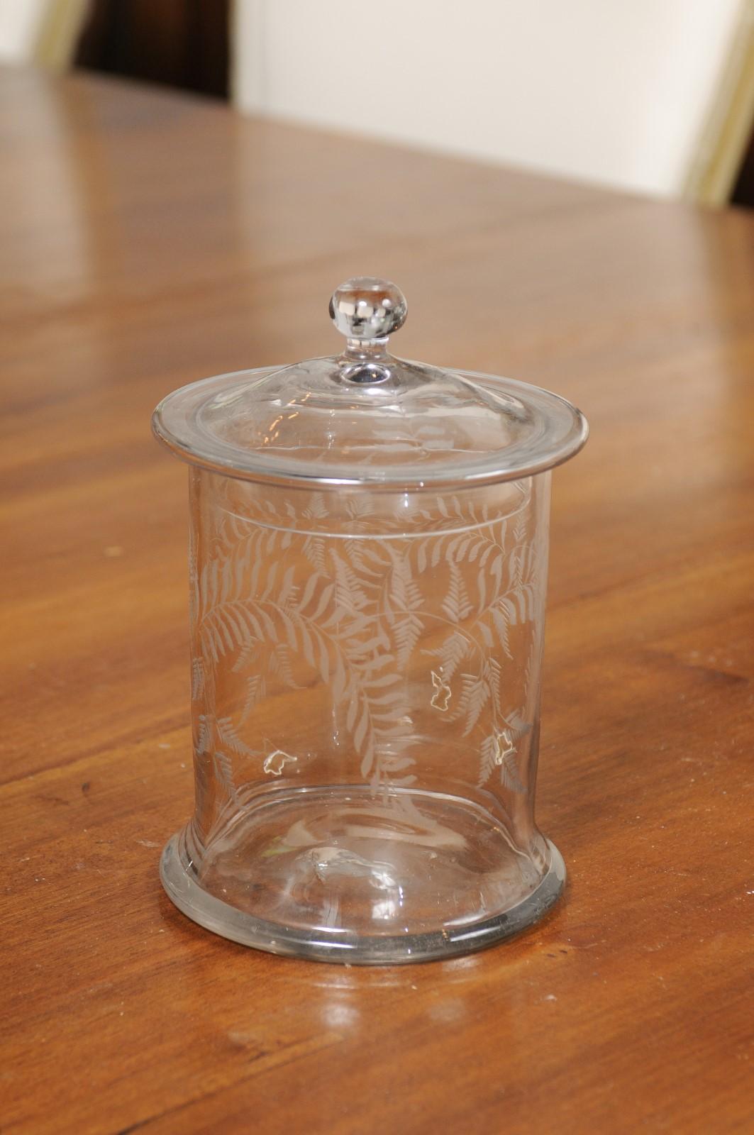 An English glass biscuit jar from the 20th century with etched foliage motifs and domed lid. Created in England during the 20th century, this glass biscuit jar features a cylindrical body adorned with fern motifs etched on the body. Topped with a