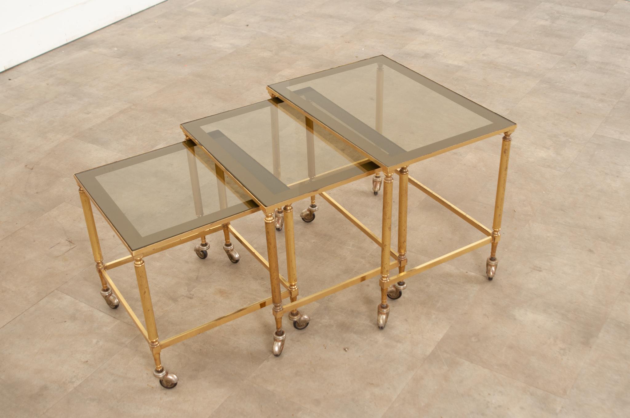 Set of three 20th century English, brass nesting tables with inset tinted glass tops supported by fluted column legs capped with turned finials and finished with casters allowing for effortless mobility. Versatile and substantial, these tables can