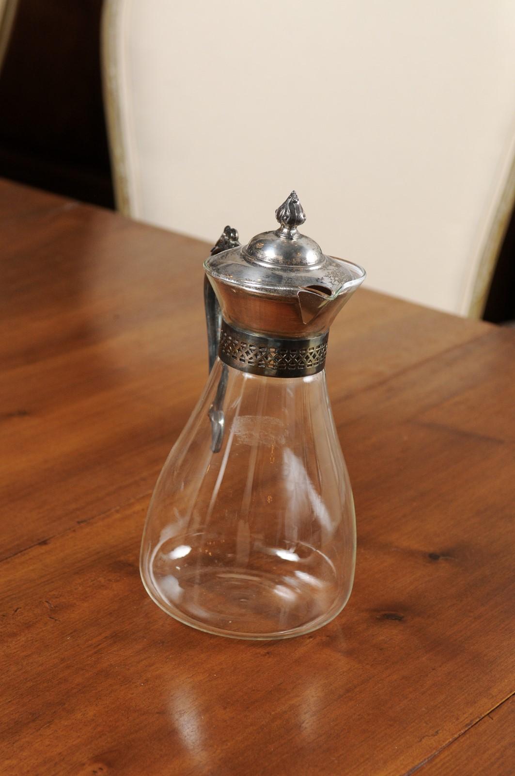 An English glass decanter from the 20th century, with silver accents. Created in England during the 20th century, this decanter features a glass body topped with a silver lid and back handle. Adorned with a flaming finial, latticed effects on the