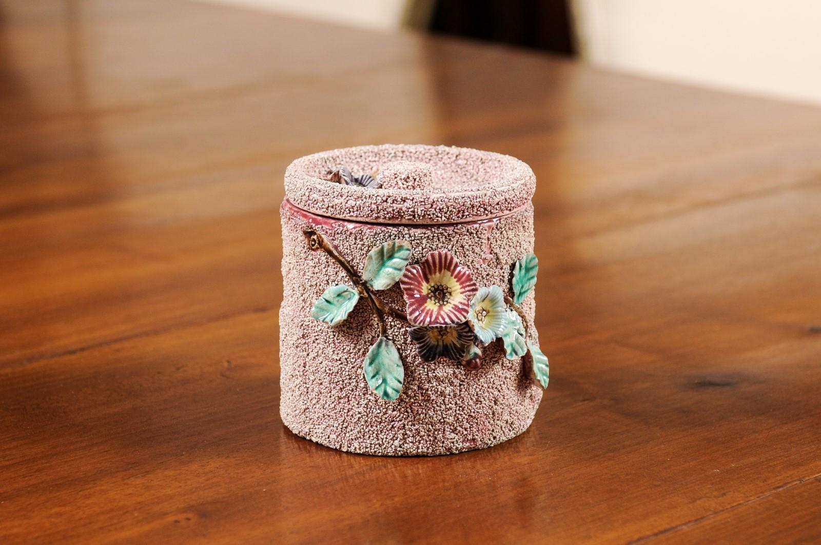 An English pottery box from the 20th century, with floral motifs, textured ground and lid. Created in England during the 20th century, this decorative container features a cylindrical body topped with a conforming lid. Adorned with red, purple and