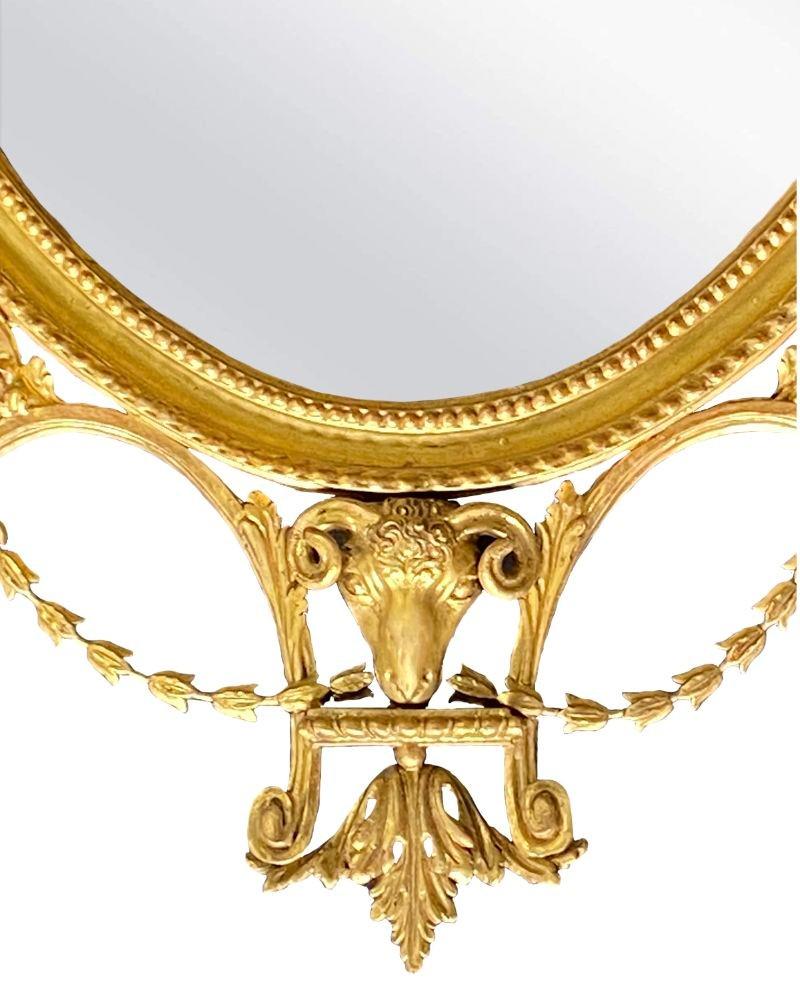 English Adams style giltwood wall mirror with delicate swags coming from the gilt ribbon design on the top of the mirror. A graceful urn is a focal point in the middle of the mirror with a ram's head adorning the base of the