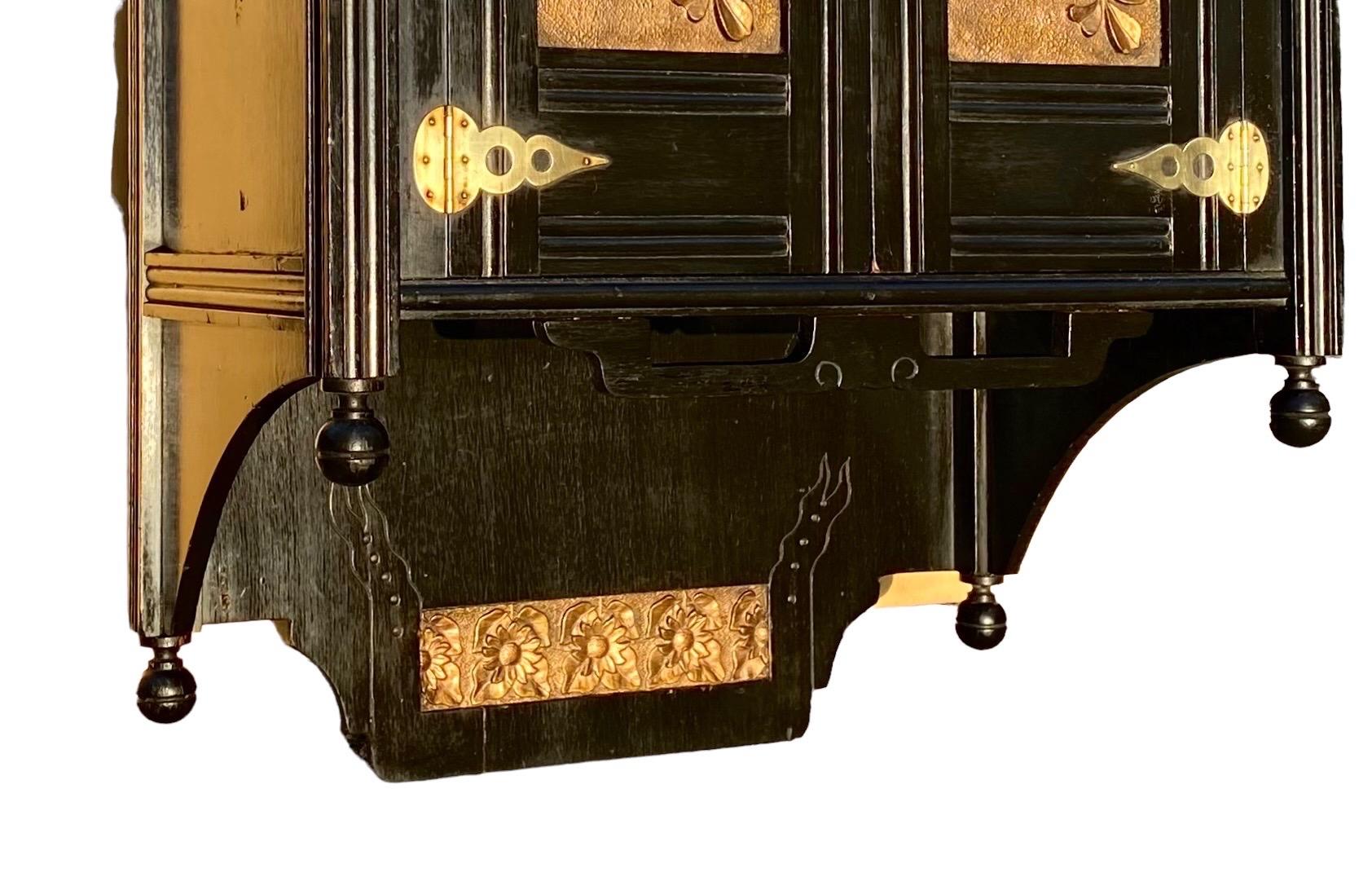 English Aesthetic Ebonized Wood and Brass Wall Cabinet, c. 1880, the finialed back splash with a relief brass floral plate, over a double door cupboard with brass floral relief panels, above a shaped shelf on a finialed bottom.

Wonderful to place
