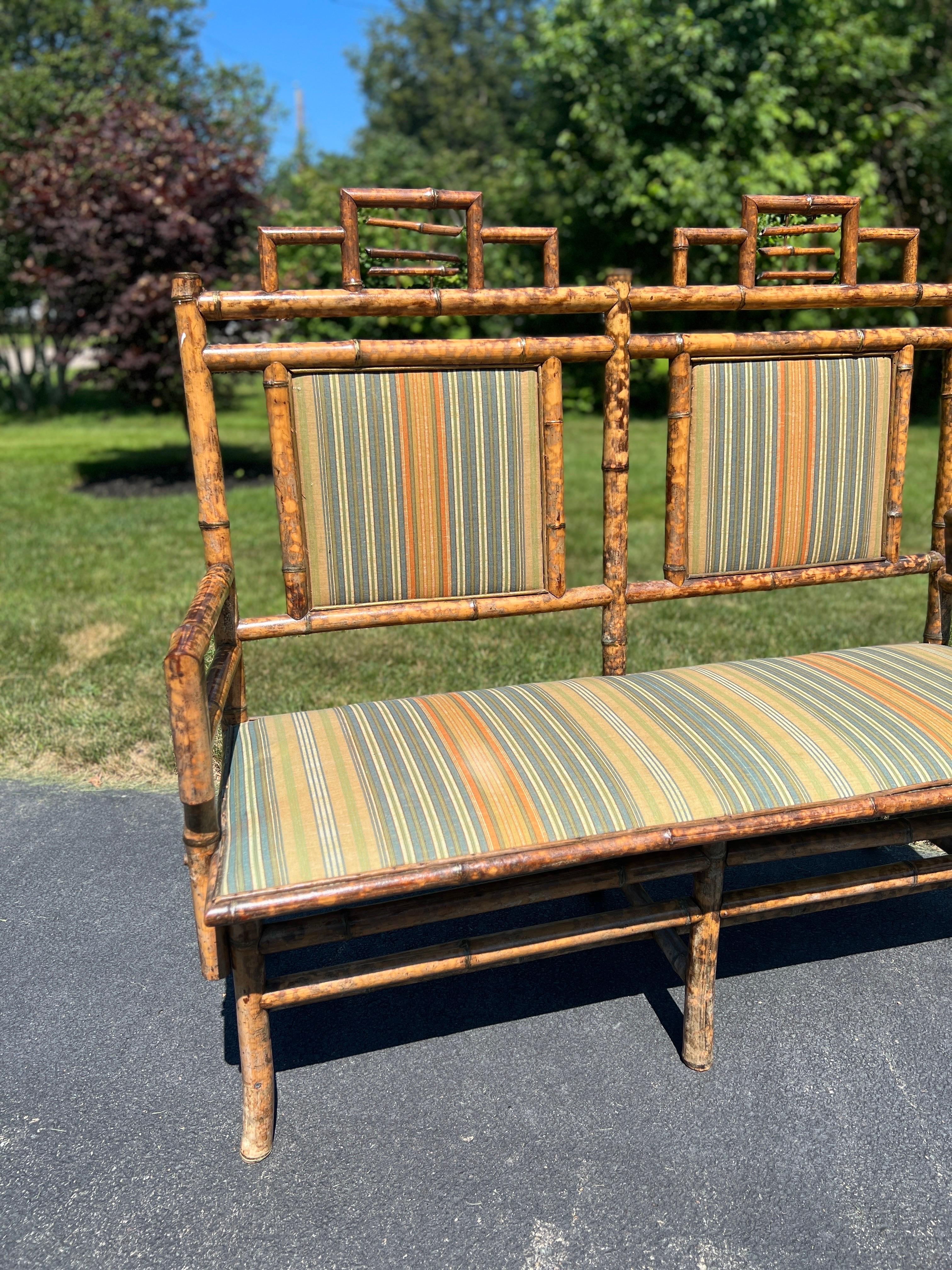 English, circa 1920. An antique English Aesthetic movement bamboo bench for two. The bench is upholstered with a traditional fabric that brings stunning lines to accent the beautiful bamboo construction on the bench. The head crest has a chinoiserie