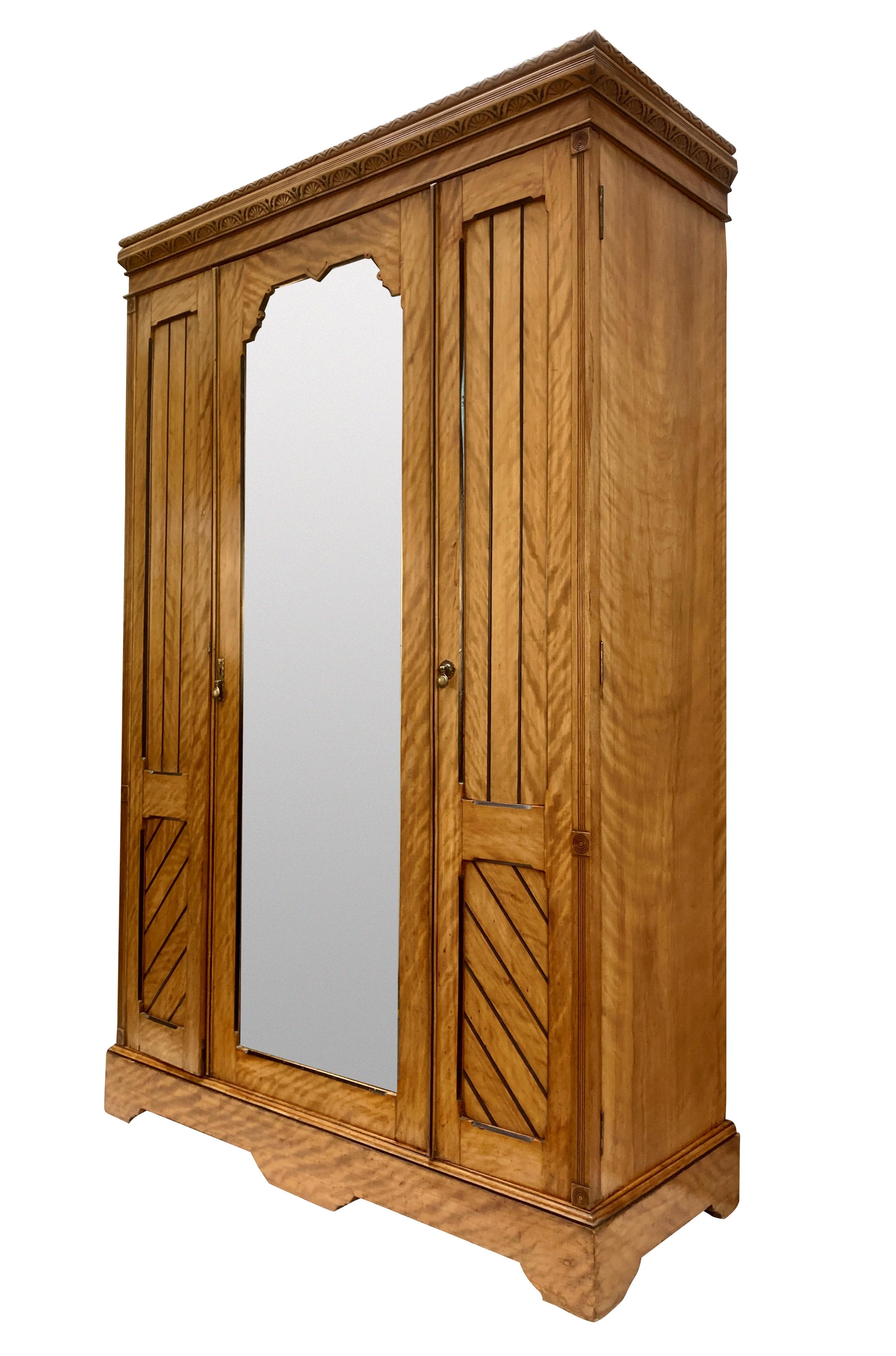 Un armadio dell'Aesthetic Movement inglese di fine Ottocento, in tre sezioni. In betulla satinata e dettagli ebanizzati, ha tre profondi cassetti e scomparti per appendere. La porta centrale con il pannello a specchio, che presenta alcuni punti di