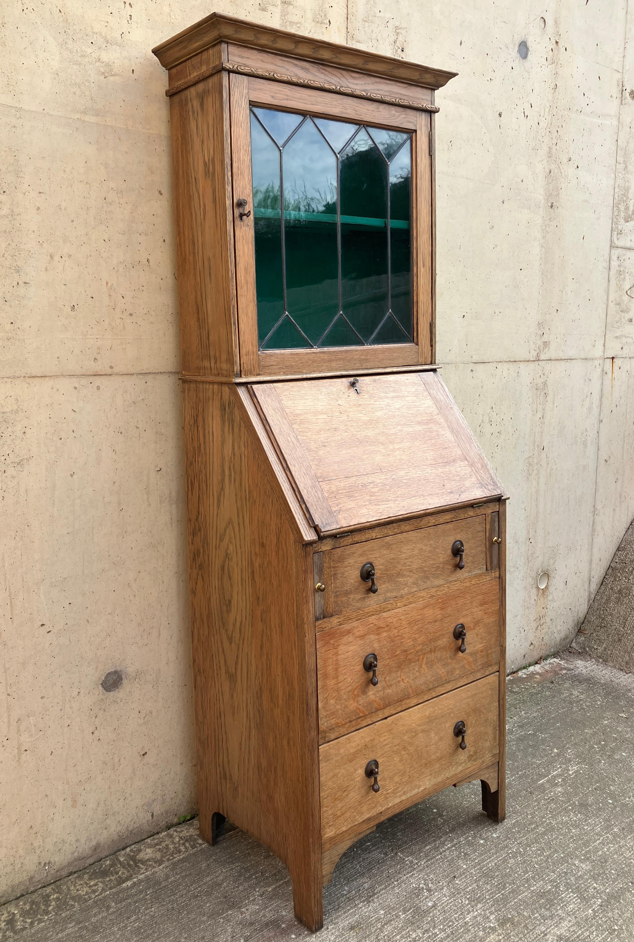 This is a beautiful compact solid oak cabinet that opens out to a bureau. The antique cabinet has a host of drawers for storage and a slim display cabinet on top with a lovely glass door with lead details. Both the vitrine on top and the bureau have