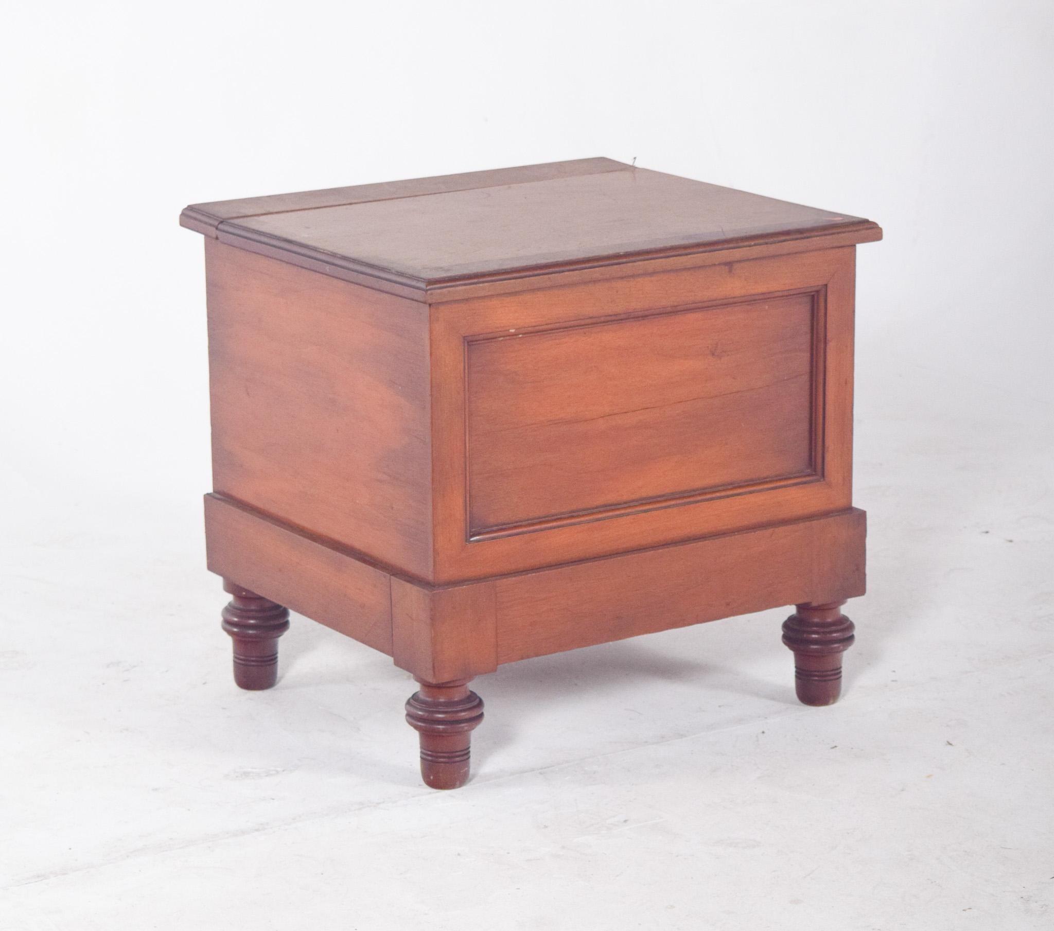 Antique wooden bedside table. Simple in shape, the top lid is hinged by metal hinges which reveal, when opened, a circular cut-out with a ceramic pot still inside. It rests on four short turned feet, typical of utilitarian period furniture,