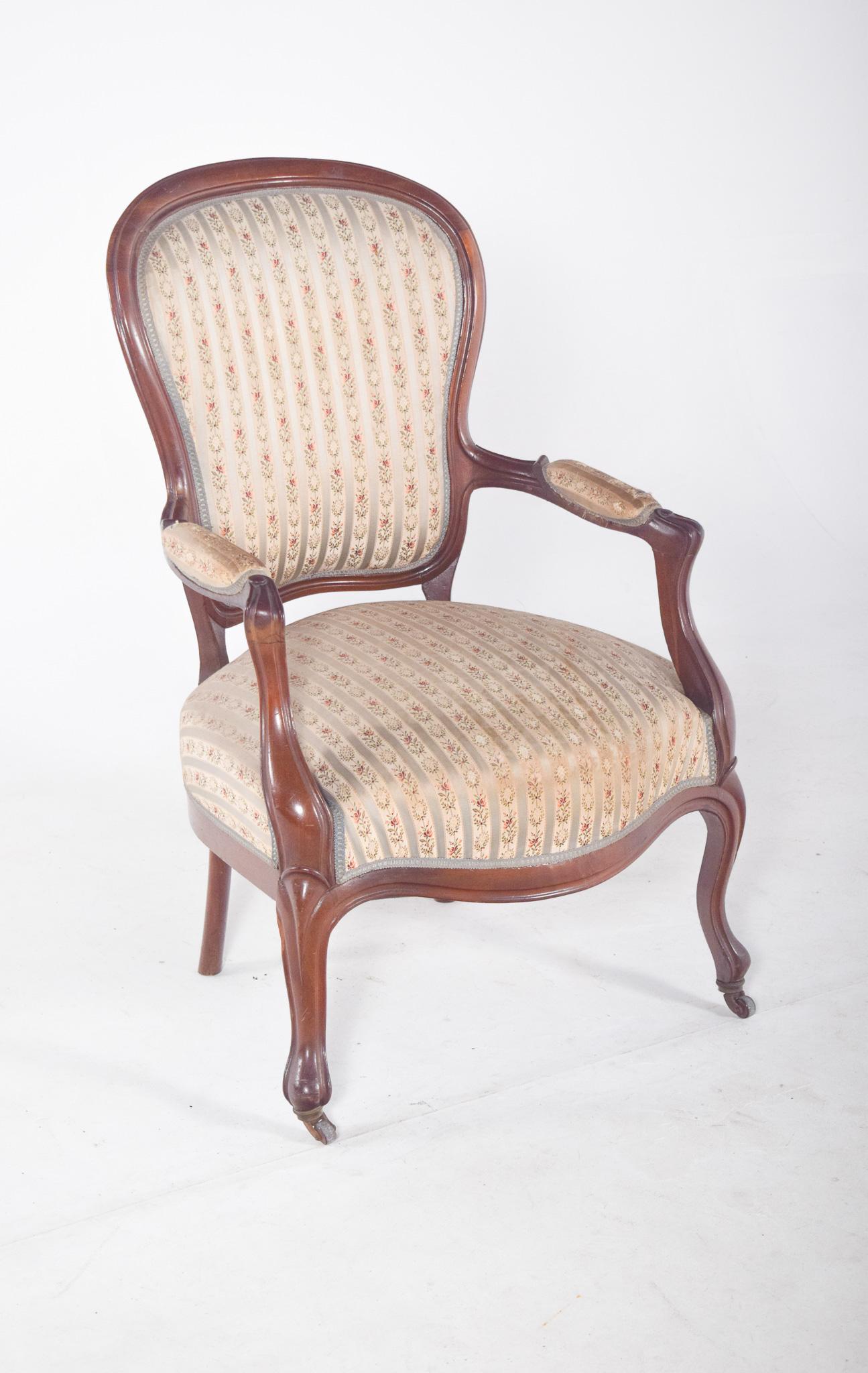 Fauteuil, tapissé en tissu à fleurs. Les formes sont arrondies, avec un dossier à cadre incurvé et des accoudoirs moulés, se terminant par des supports rembourrés. Les pieds avant sont arqués, avec de petites roues en bois à la base, tandis que les