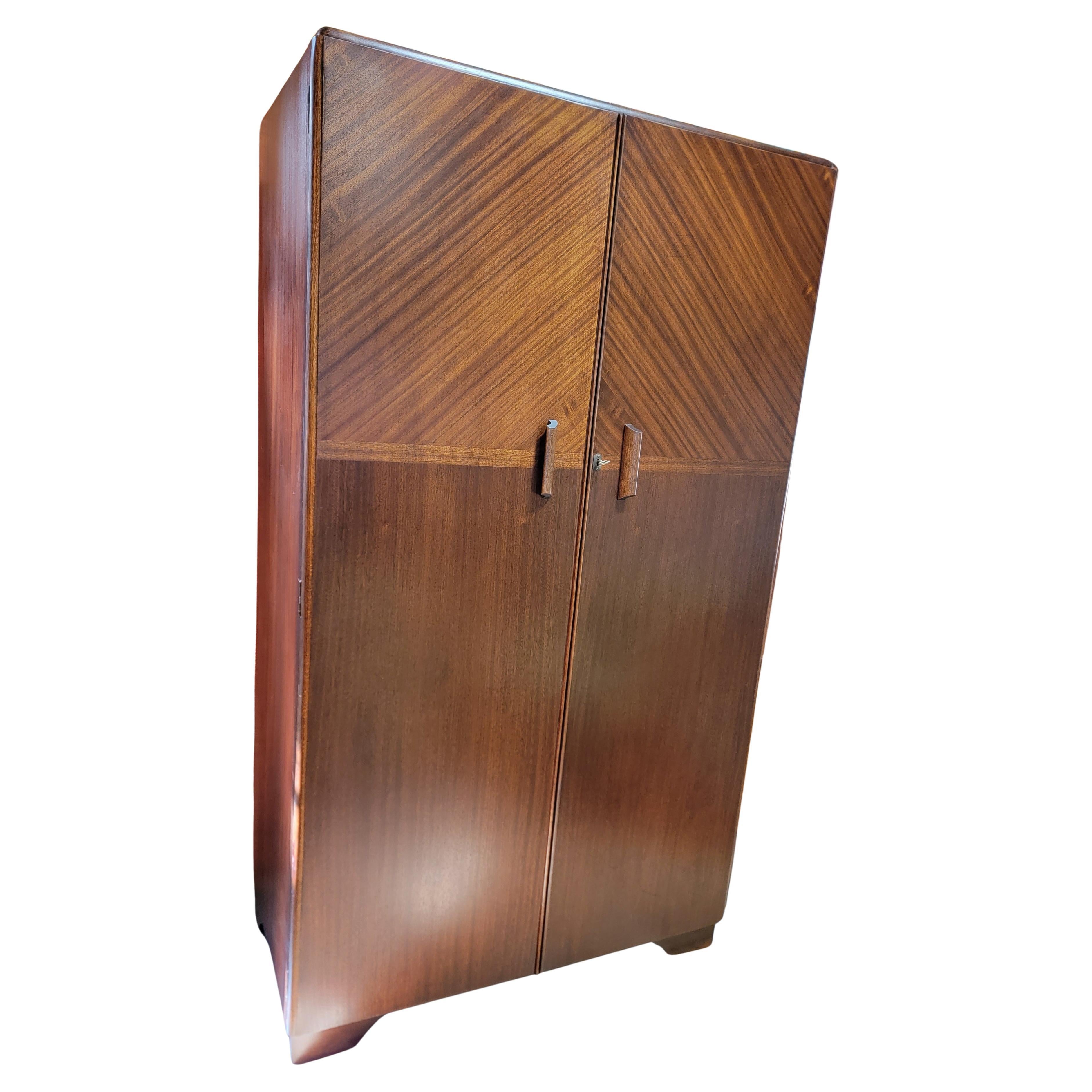 An English Art Deco double door armoire / wardrobe with a beautiful book-matched ribbon cut mahogany detail. Interior fully compartmentalized with various drawers, clothes hanger and mirror. All compartments retain original labeling to assist with