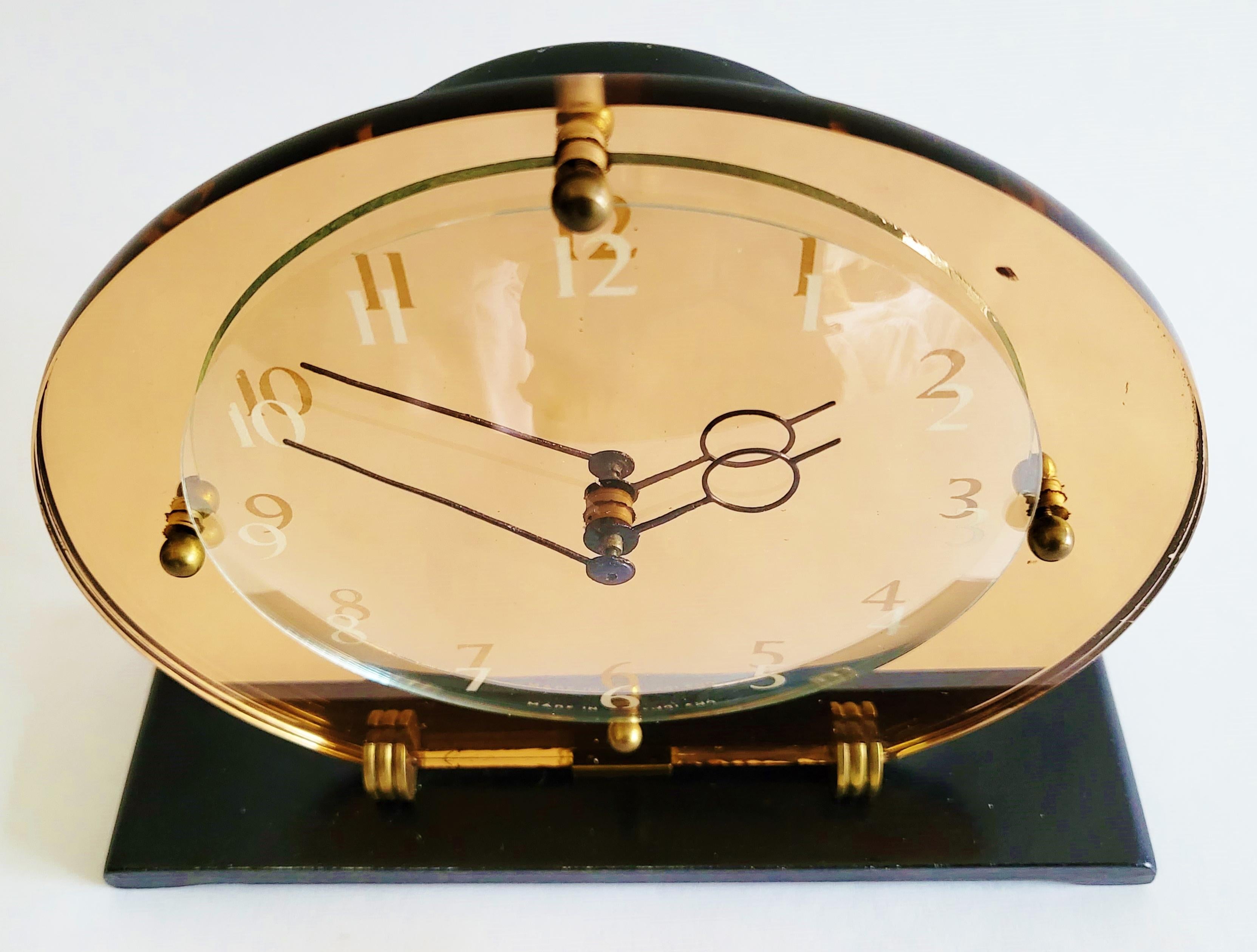 This stylish English Art Deco mechanical clock features an offset peach mirror backplate supported by two ridged brass disc feet that are mounted together with a central brass support to a black lacquered oblong base with chamfered ends. The white
