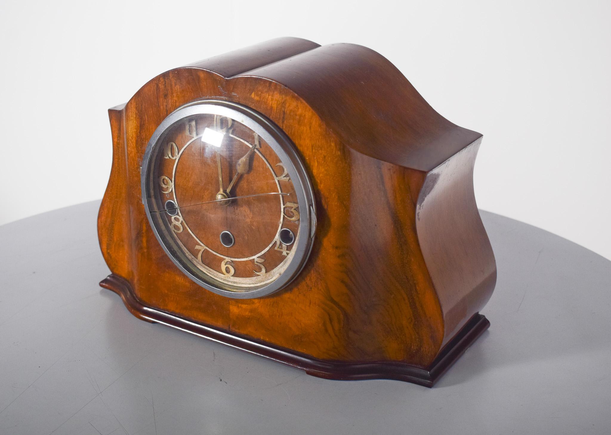 Art Deco table clock. It has an elegant design with a carved wooden case and a varnished finish, emphasising the natural tone of the wood. Its curved, symmetrical shape gives it a classic charm. The circular dial features embossed gold numerals,