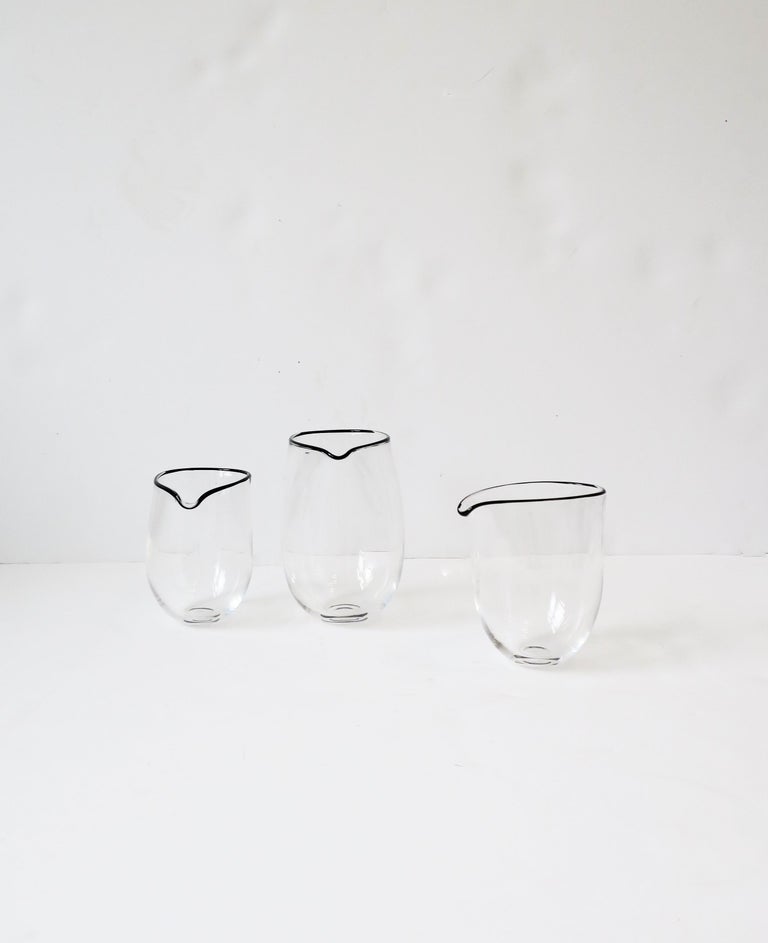 A beautiful hand-made set of three (3) modern/minimalist style art glass beaker pitcher cocktail vessels, made in England by artist designer Simon Moore, circa late-20th century, 1990s. Each, hand-crafted, are transparent crystal with black art
