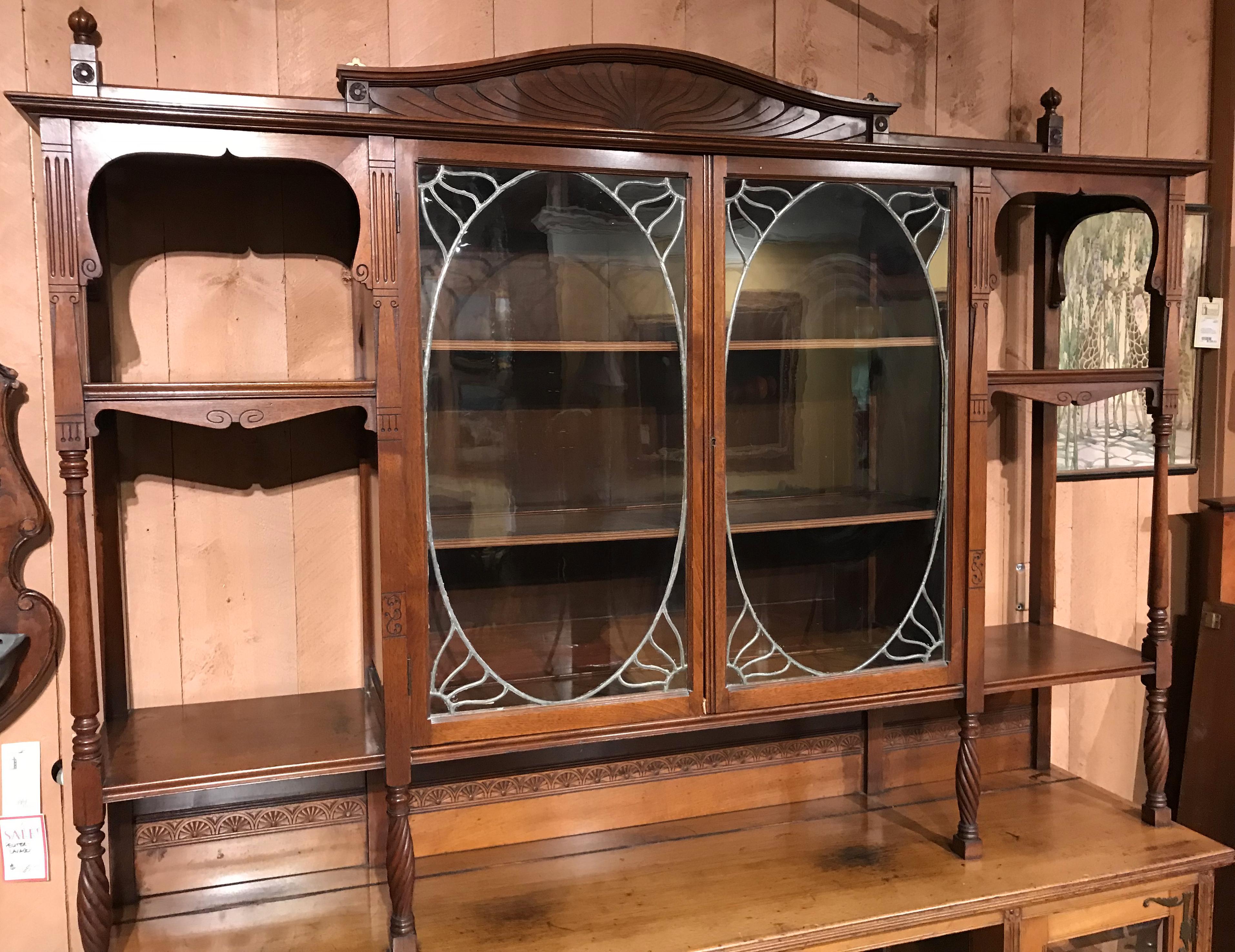 A fine two part English Art Nouveau étagère or bookcase, its upper case featuring a radial carved crest and spiral turned finials surmounting a center cabinet with leaded glass doors flanked by open shelves accented with carved rosettes and turned