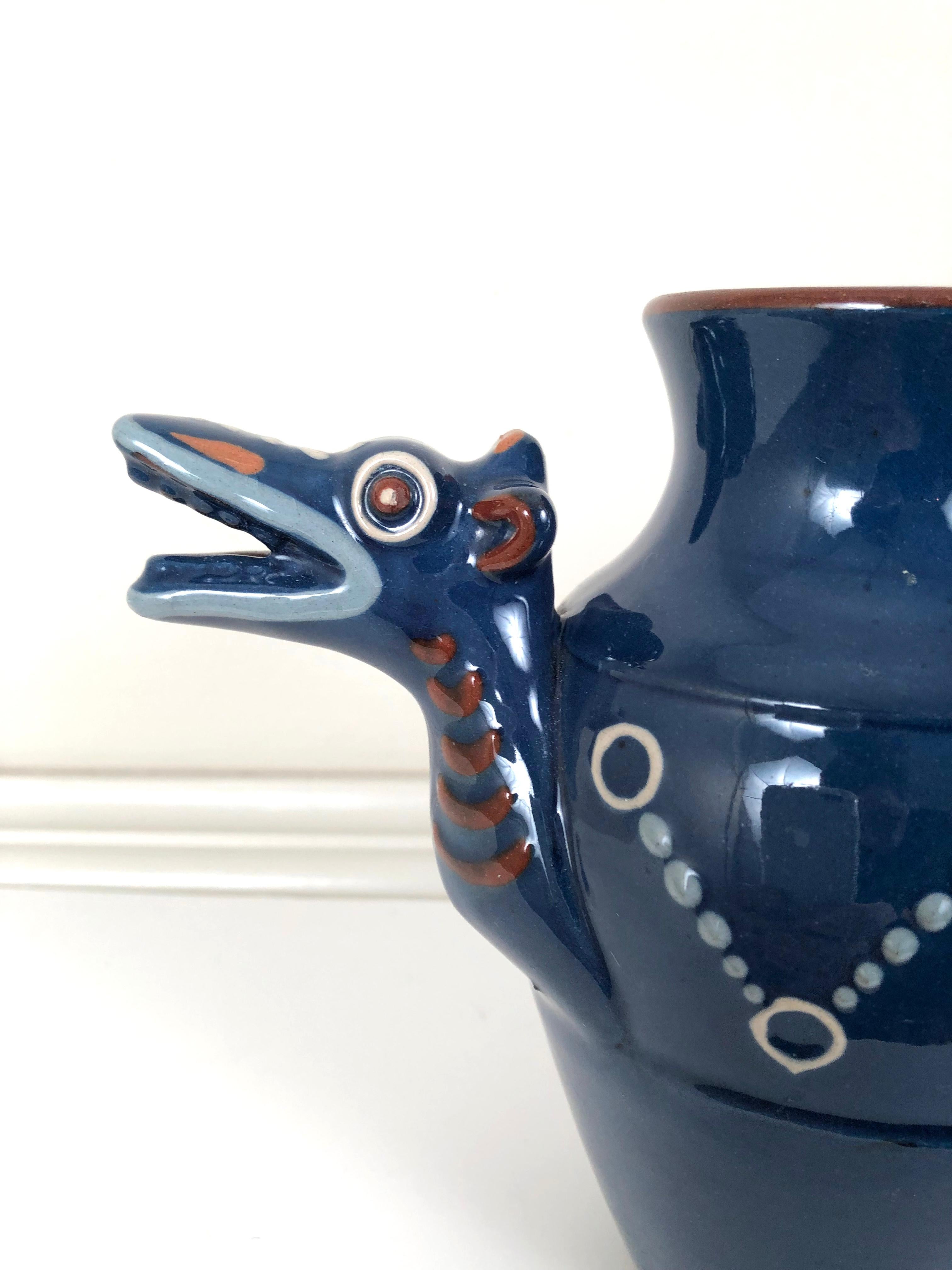 An English Arts & Crafts period pottery Longpark dragon form pitcher, the design attributed to Blanche Vulliamy, its head forming the spout and the curled tail with fins forming the handle, slip decorated with circular motifs in light blue, cream