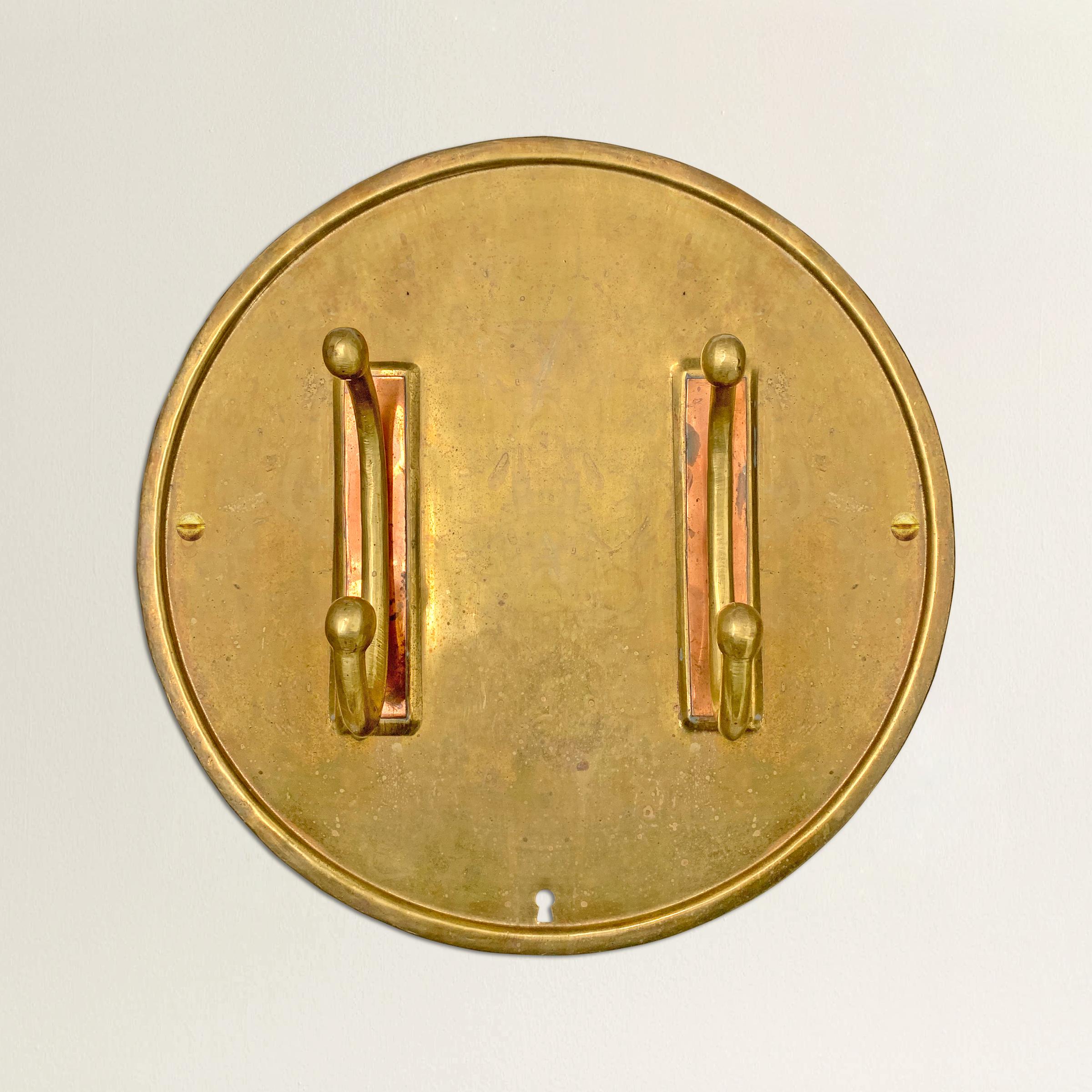 An über chic early 20th century English Arts & Crafts brass and copper coat rack with two hooks with copper backplates attached to a large round brass backplate, and a curious little cutout key hole at the bottom. We've left the finish untouched,