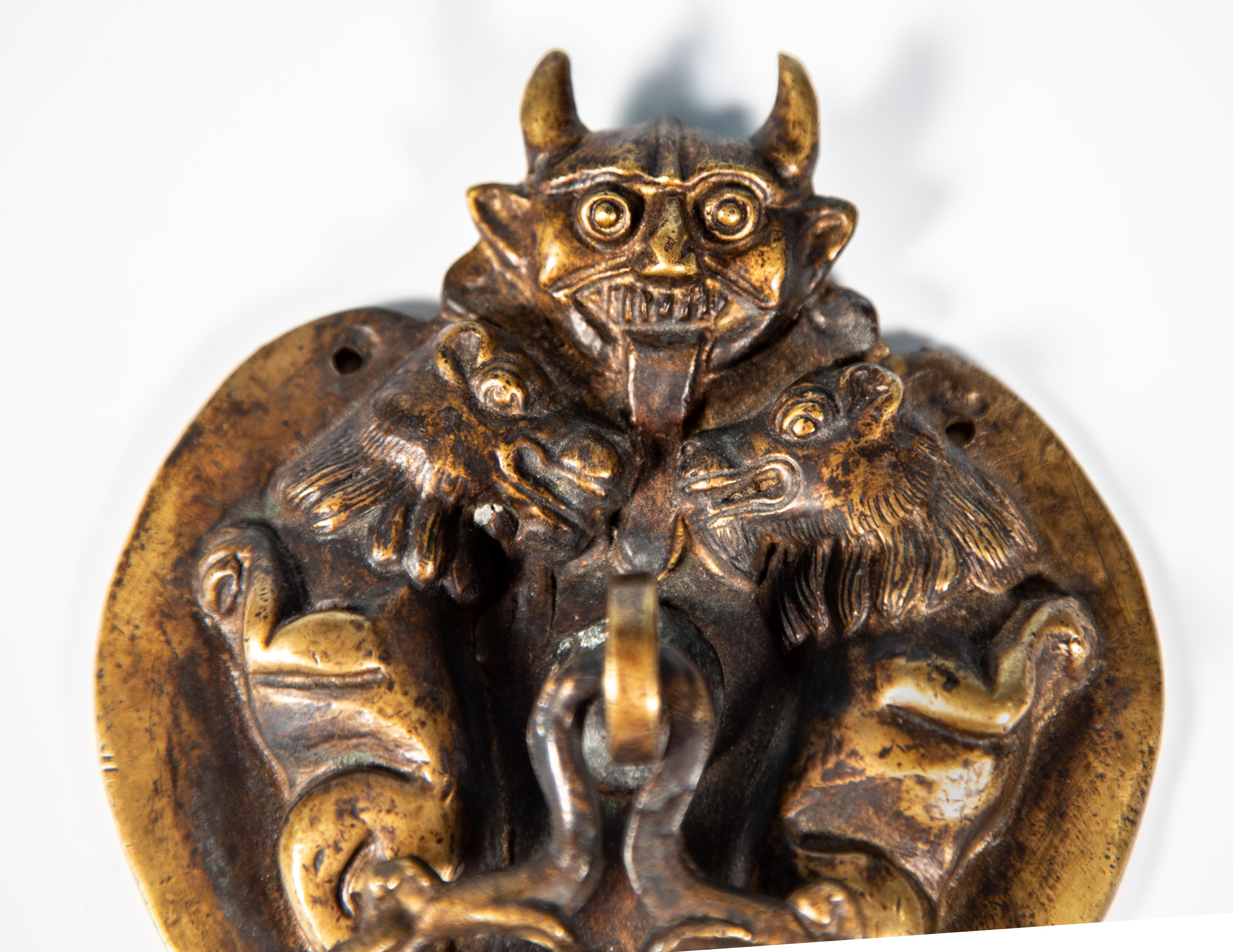 English Arts & Crafts cast bronze devil door knocker circa 1900. Front on image of a devil sticking out his tongue flanked by two guardian lions. Knocker hanging from the enter position. Good art casting and shows modeling from the back. Hand chased