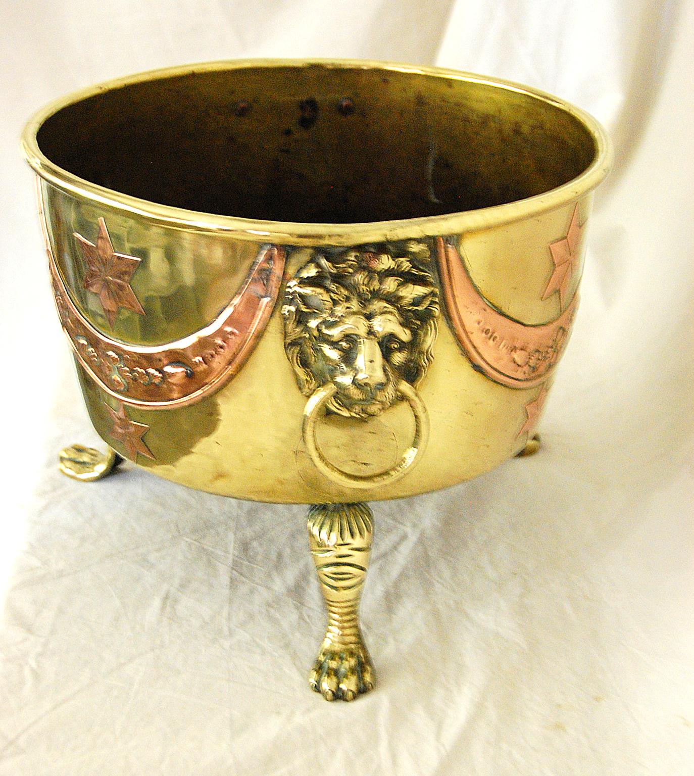 English Arts & Crafts period 19th century brass and copper footed jardinière or log bin. This heavy quality unusual brass footed jardinière is overlaid with swags and stars of repousse copper and has bold lion ring handles.