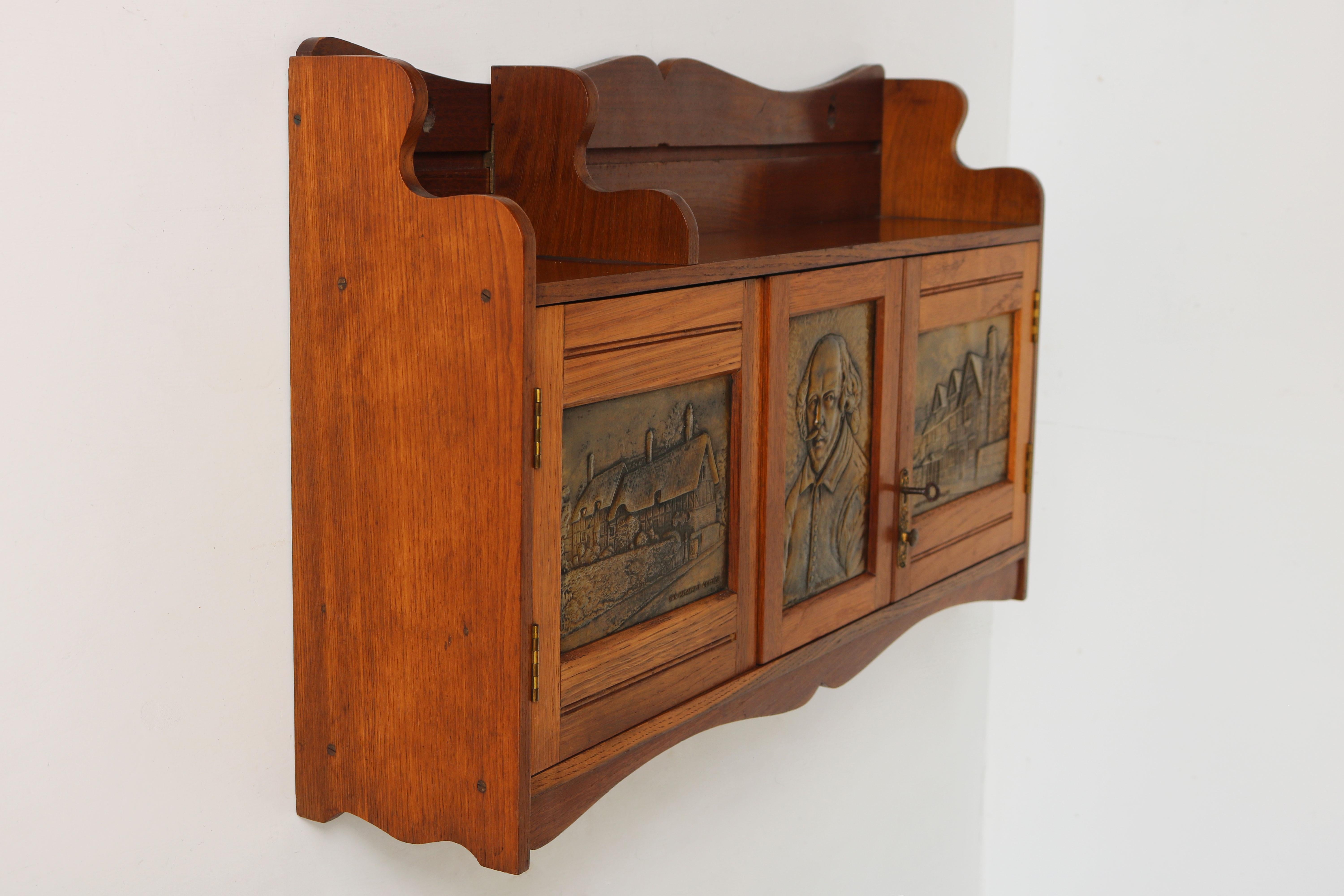 Lovely English Arts & crafts wall cabinet with brass panels displaying Shakespeare made from oak. 
The cabinet has many functions: on top is a adjustable bookshelf. The right door opens using a lock & key. Behind this door is a writing compartment