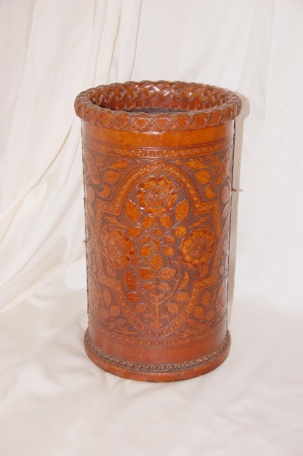English Arts & Crafts hand embossed leather (cowhide) waste basket or stickstand. The elegant leather hand tooling has a floral motif within curving borders. The base has brass tacks, the upper rim is woven leather and the side seams are braided