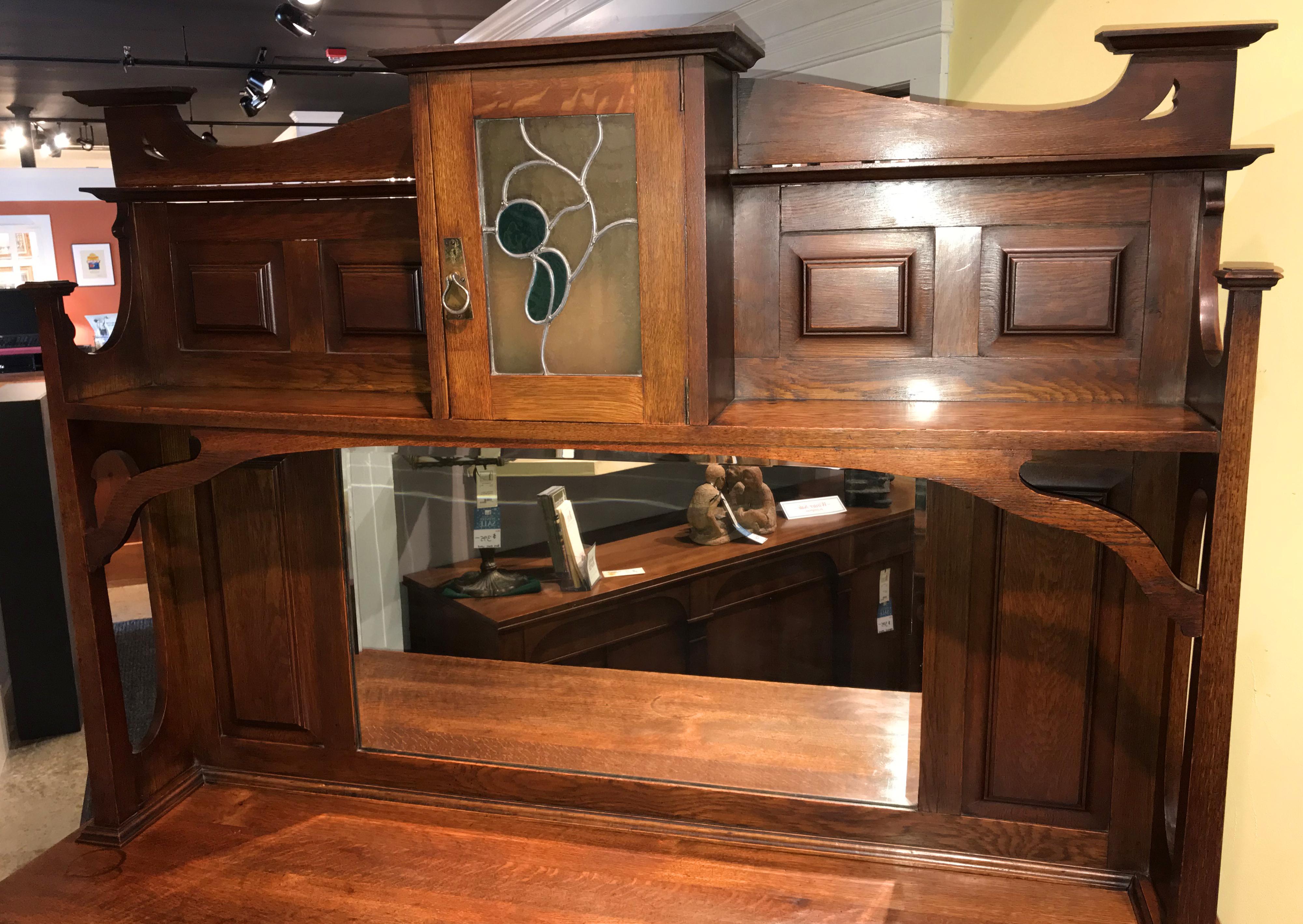 A wonderful English Arts & Crafts possibly by Liberty & Company two-piece oak sideboard with mirrored upper section featuring a centre upper shelf door with stained leaded glass, foliate decorated, which opens to a small storage compartment, flanked