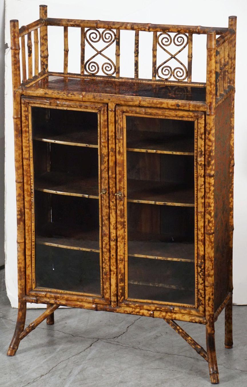 A handsome tall English bamboo bookcase from the Aesthetic Movement period, featuring a decorative gallery top of bamboo with japan-lacquered panel, over two framed-glass doors with brass hardware, opening to an interior of three bamboo-edged