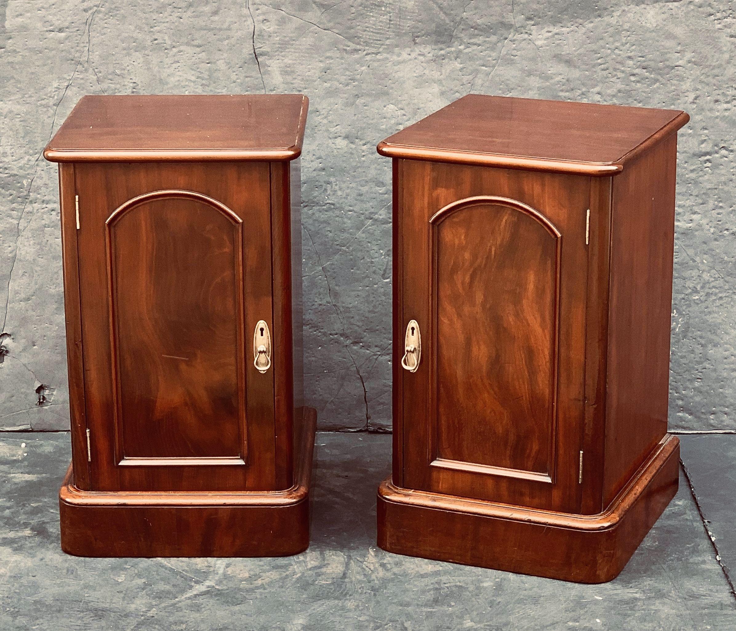 A handsome pair of English bedside chests or cabinet nightstands of mahogany - each chest with a moulded top over a cabinet door with brass escutcheon, opening to a cupboard with drawer and two shelves, set upon a plinth base.

Priced as a pair -