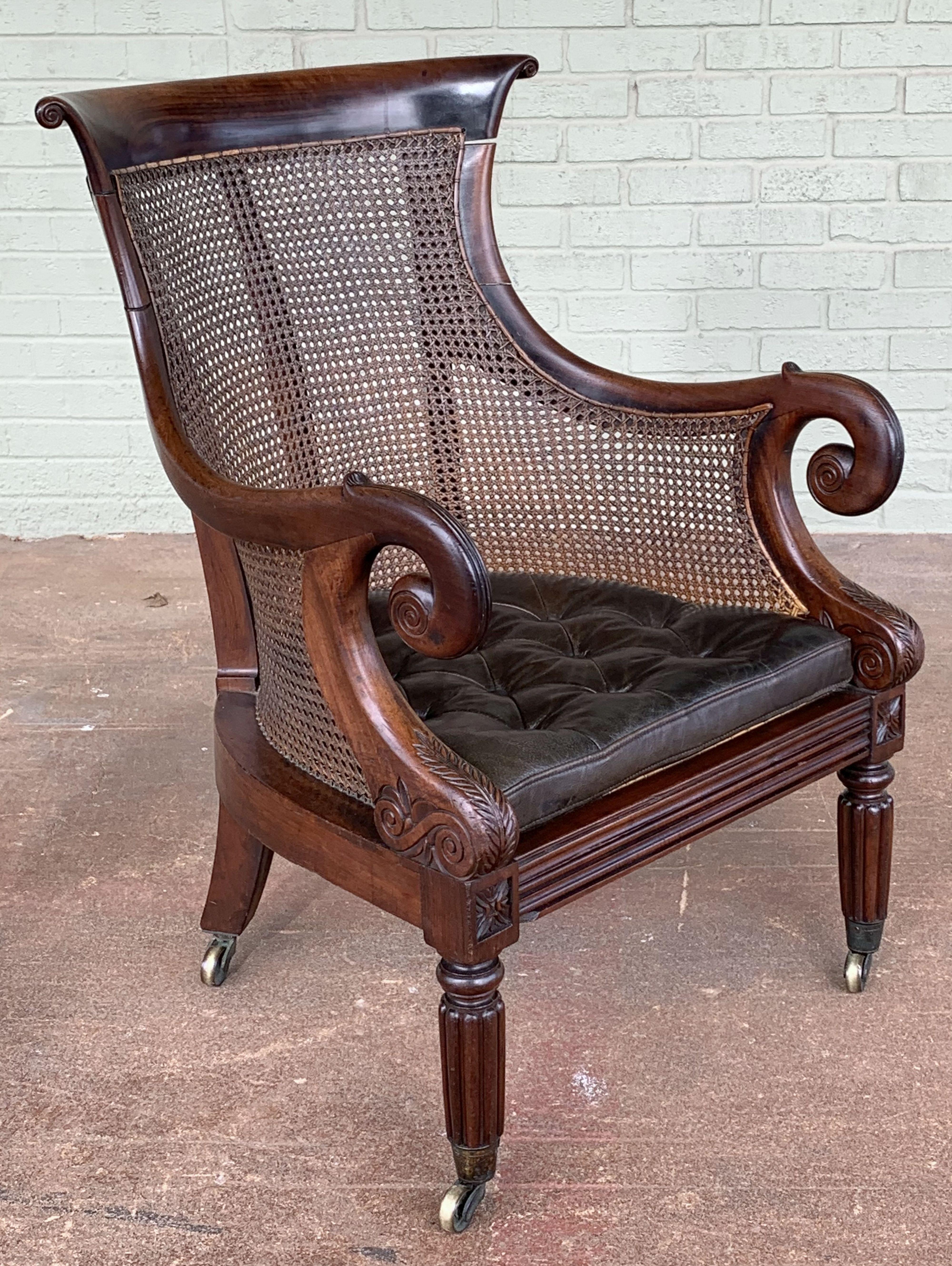 A fine, comfortable English bergère armchair or library chair of caned mahogany from the Regency Period, featuring a Classic, stylish turned scroll back and arms, removable tufted-leather cushion, on turned leg supports, with rolling brass