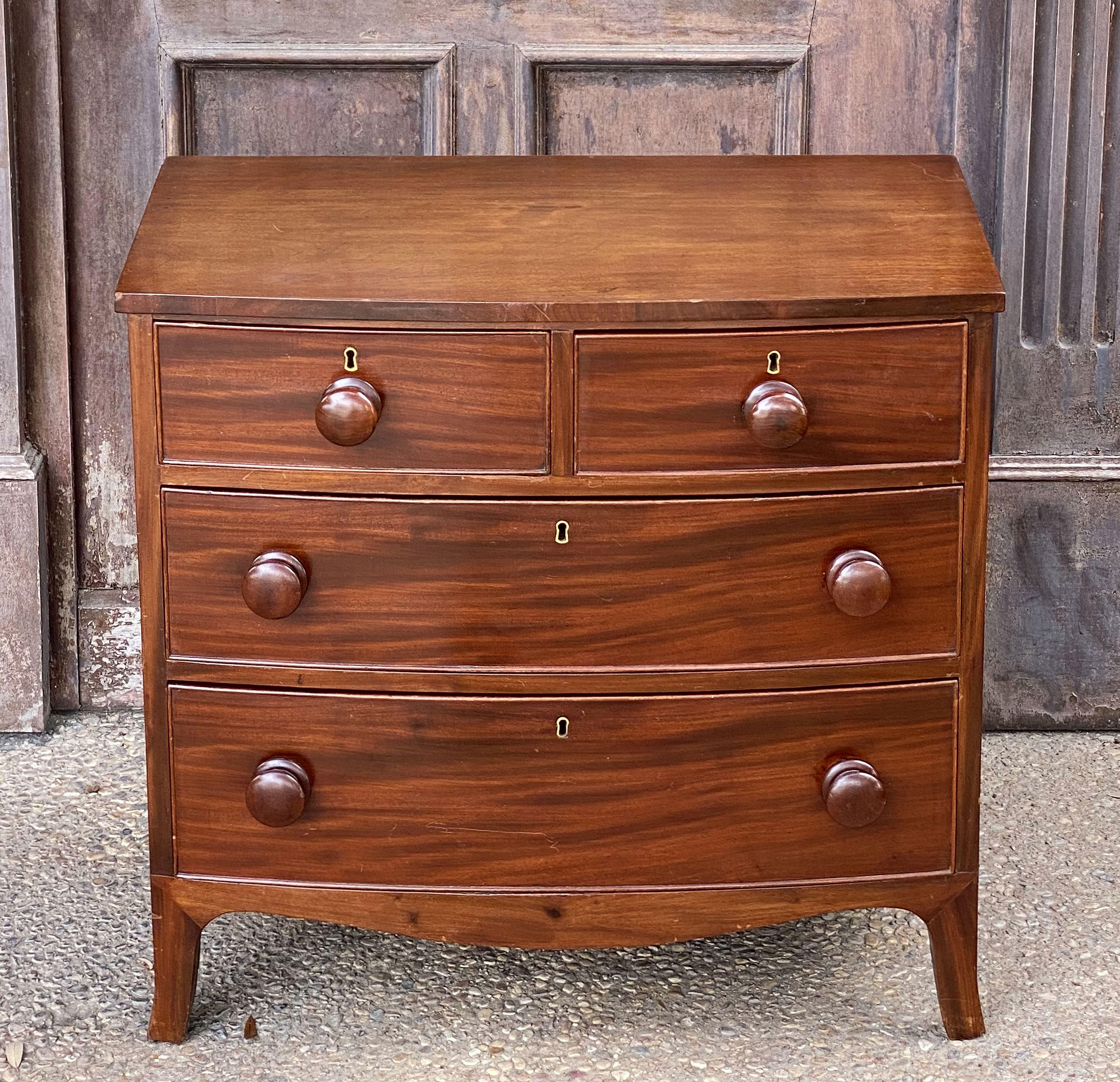 A handsome English bow-front small chest of drawers of mahogany from the 19th century - featuring a bowed top over a frieze of two small beaded drawers and two long beaded drawers, each drawer with fine mahogany veneers, knob pulls, and brass