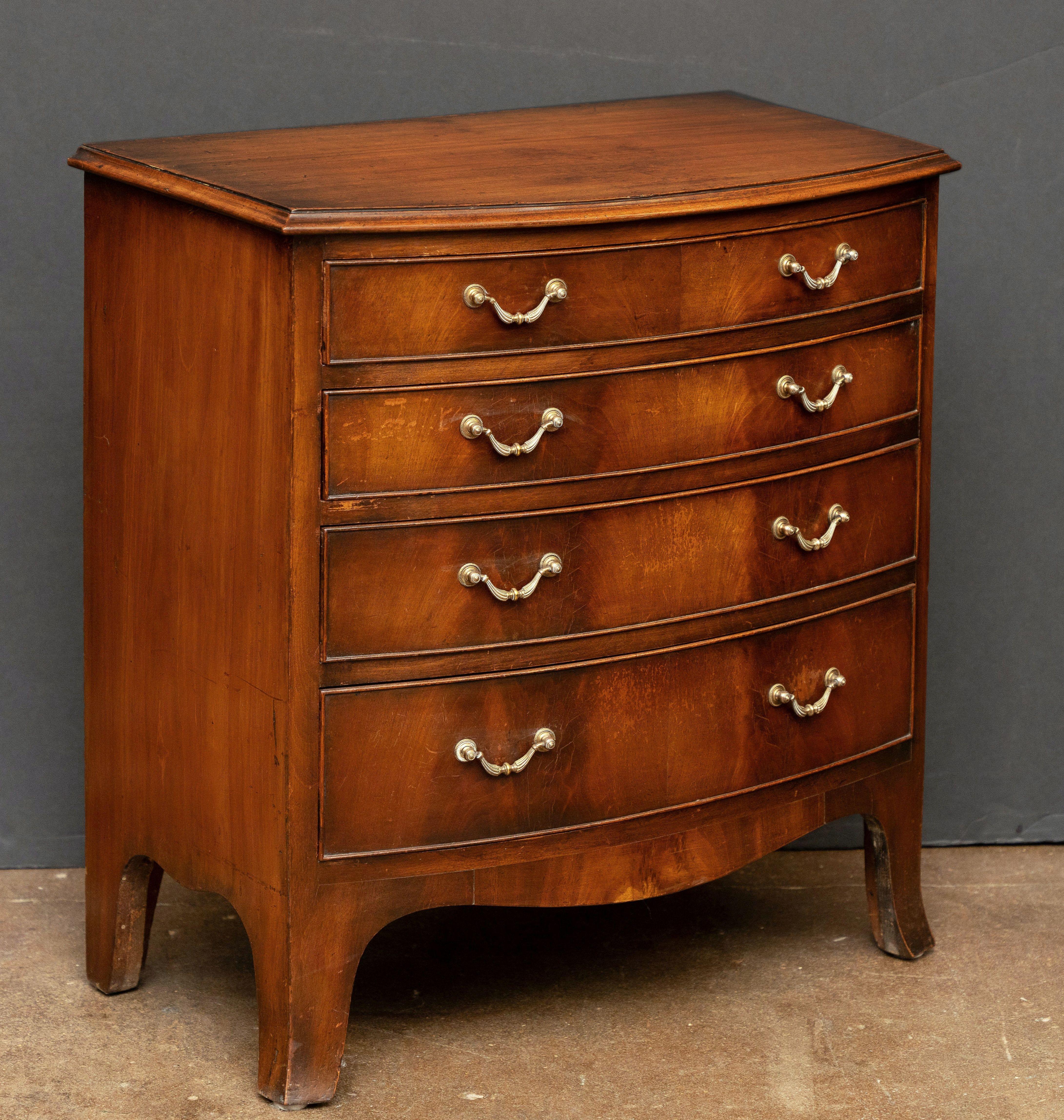 A handsome English bow-fronted small chest of mahogany from the Edwardian era, featuring a moulded top over a frieze of four drawers, each beaded edge drawer with flame mahogany veneers over an oak interior, with original reeded brass drop handles,